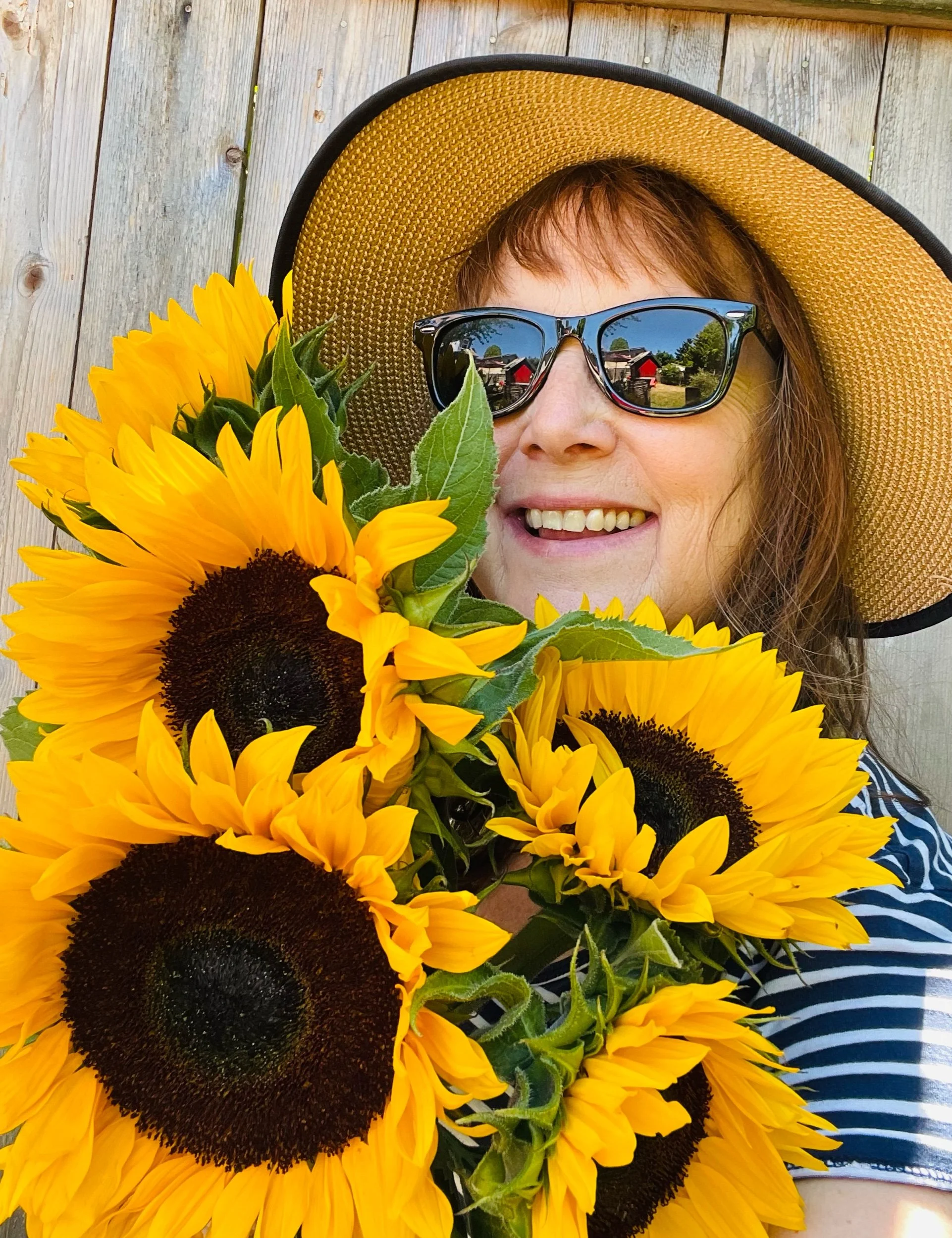 Person smiling, wearing sunglasses and a wide-brimmed straw hat, holding a bouquet of large sunflowers with dark centers, in front of a wooden background.