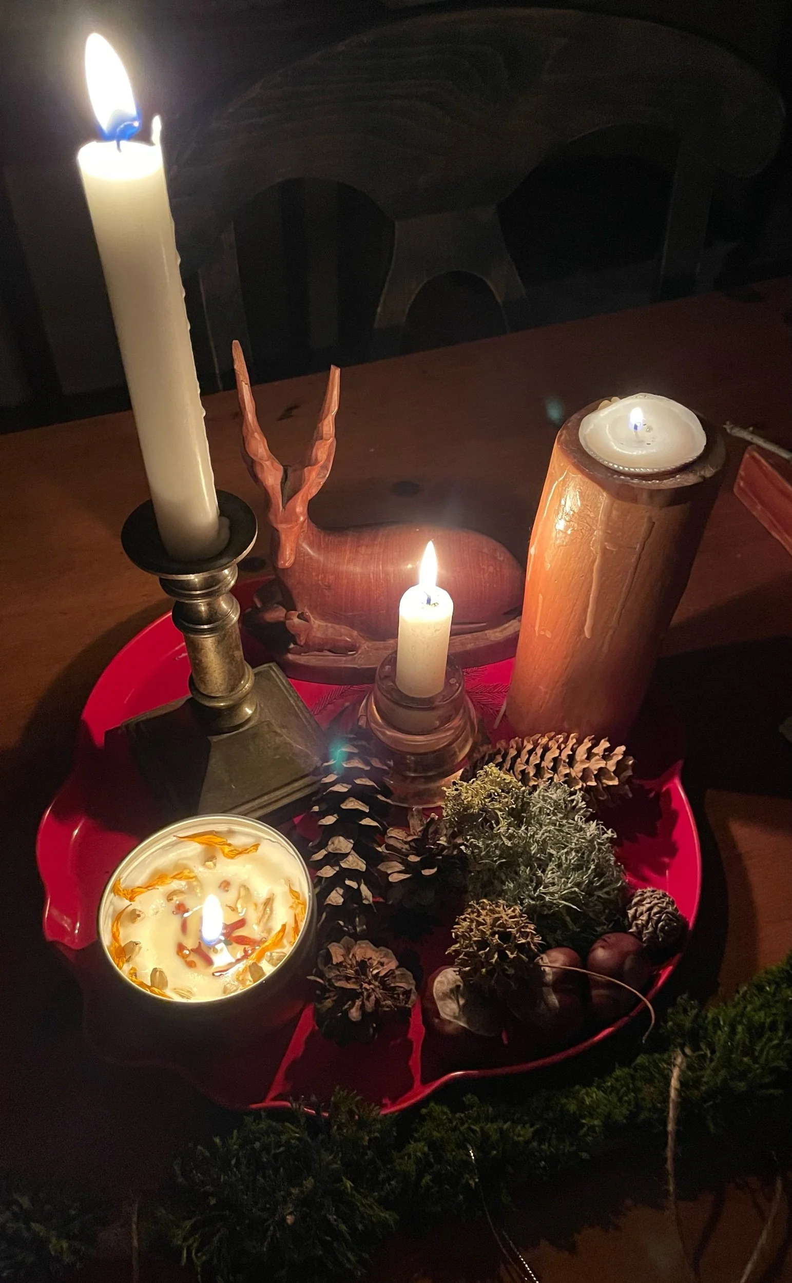 A wooden table with candles, pinecones, dried flowers, and greenery on a red tray, illuminated by candlelight, creating a cozy and festive atmosphere.