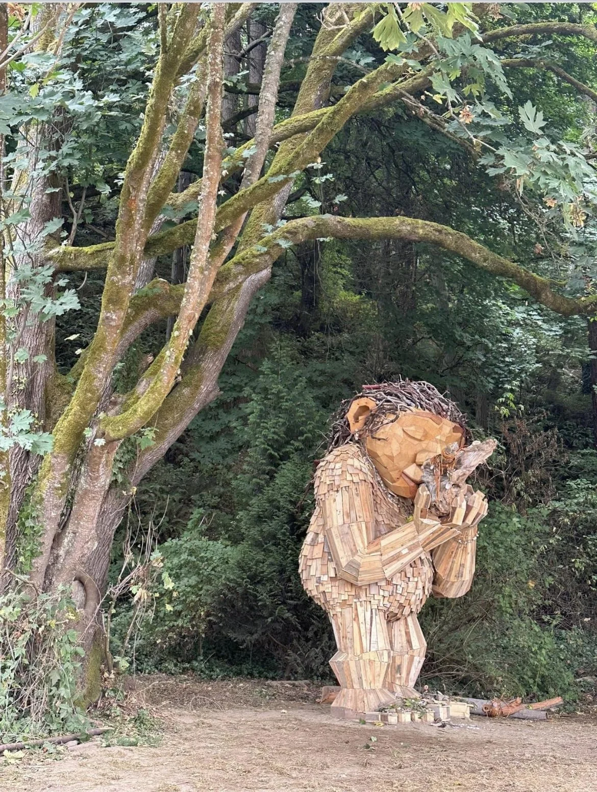 A large wooden sculpture of a child with closed eyes, sitting with knees pulled to chest and hands covering ears, made from small wooden blocks, placed outdoors among trees and greenery.