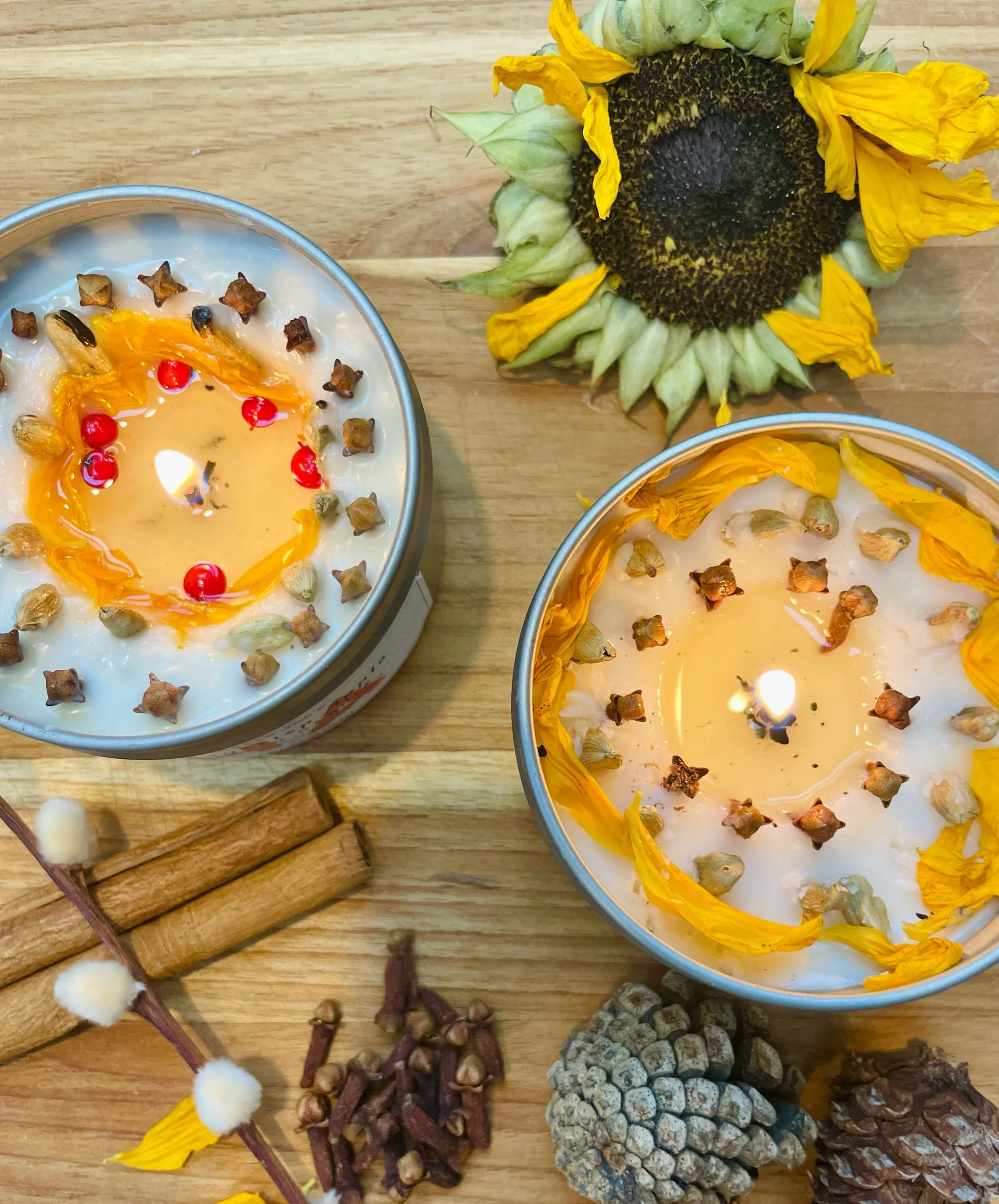 A top view of two candles decorated with dried flowers and red berries, placed on a wooden surface alongside dried pinecones, cinnamon sticks, and a sprig of artificial pussy willow.