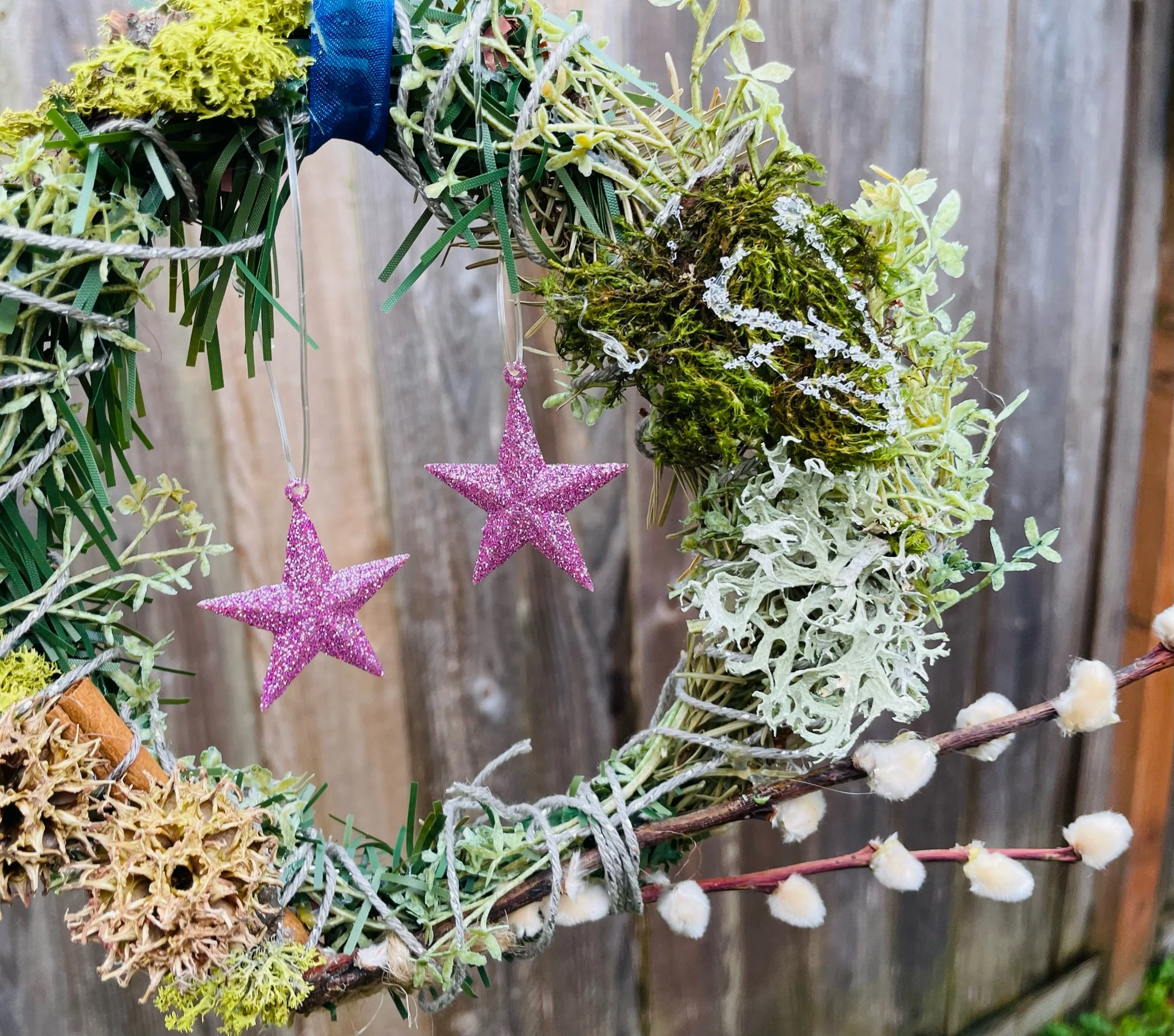 Close-up of a decorative wreath made of various green mosses, plants, and branches, with pink glittery star ornaments hanging from the wreath against a wooden background.