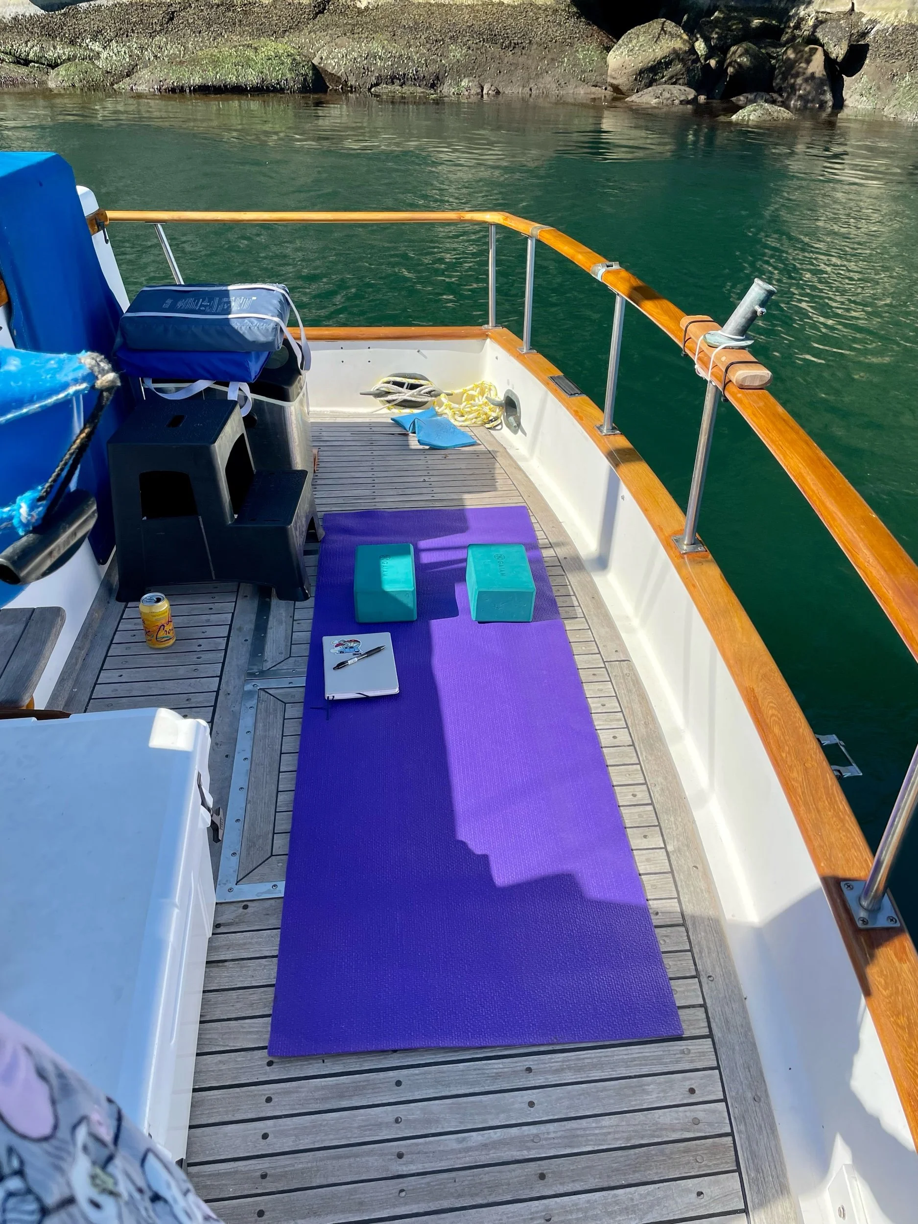 View of a boat deck with a purple yoga mat, two yoga blocks, a notebook, a pen, a water bottle, and some miscellaneous items, along with water and rocky shoreline in the background.