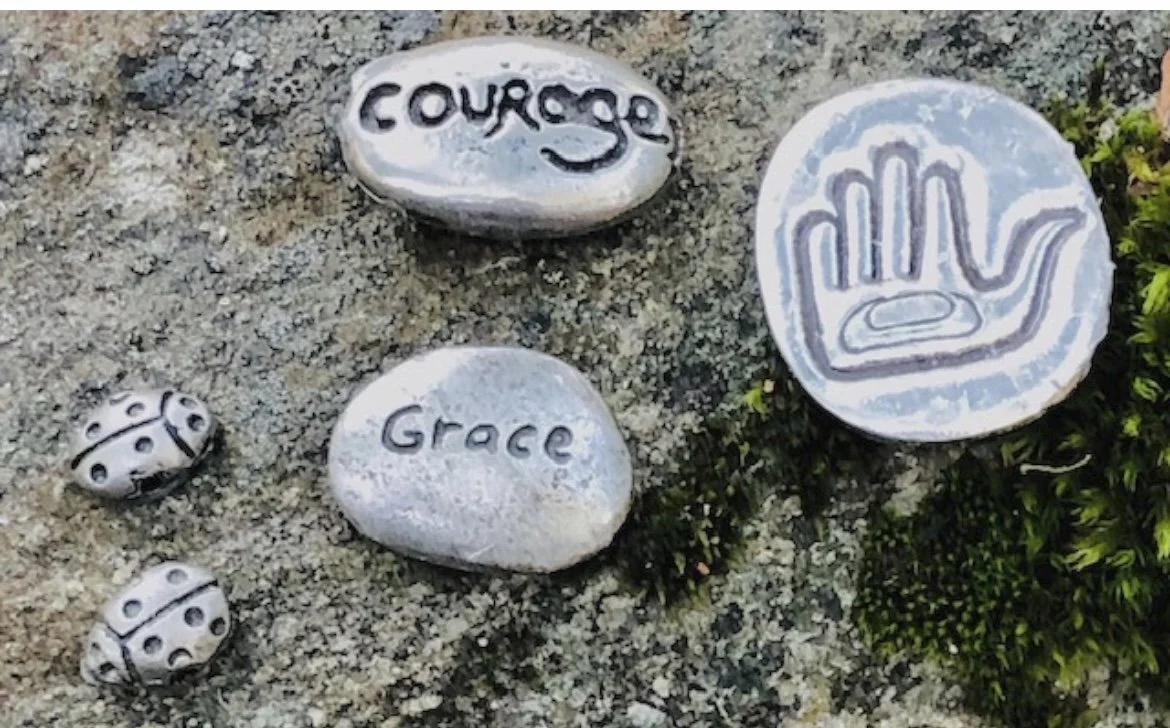 Decorative rocks on sand with the words 'Courage' and 'Grace' engraved, and a circle with a hand symbol.