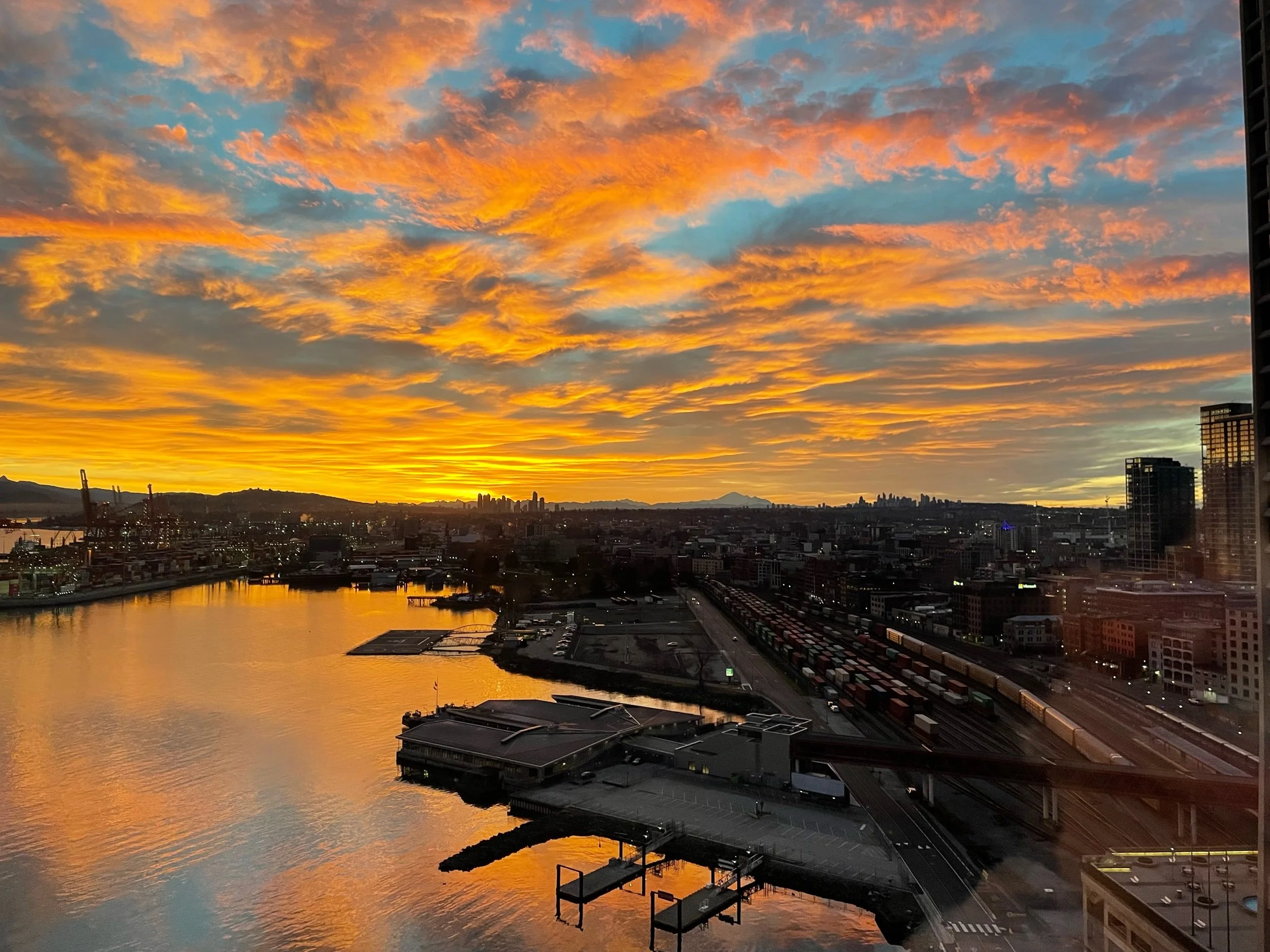 A city skyline during sunset with orange and blue clouds, a body of water reflecting the sky, and buildings including a tall tower and industrial structures in the foreground.