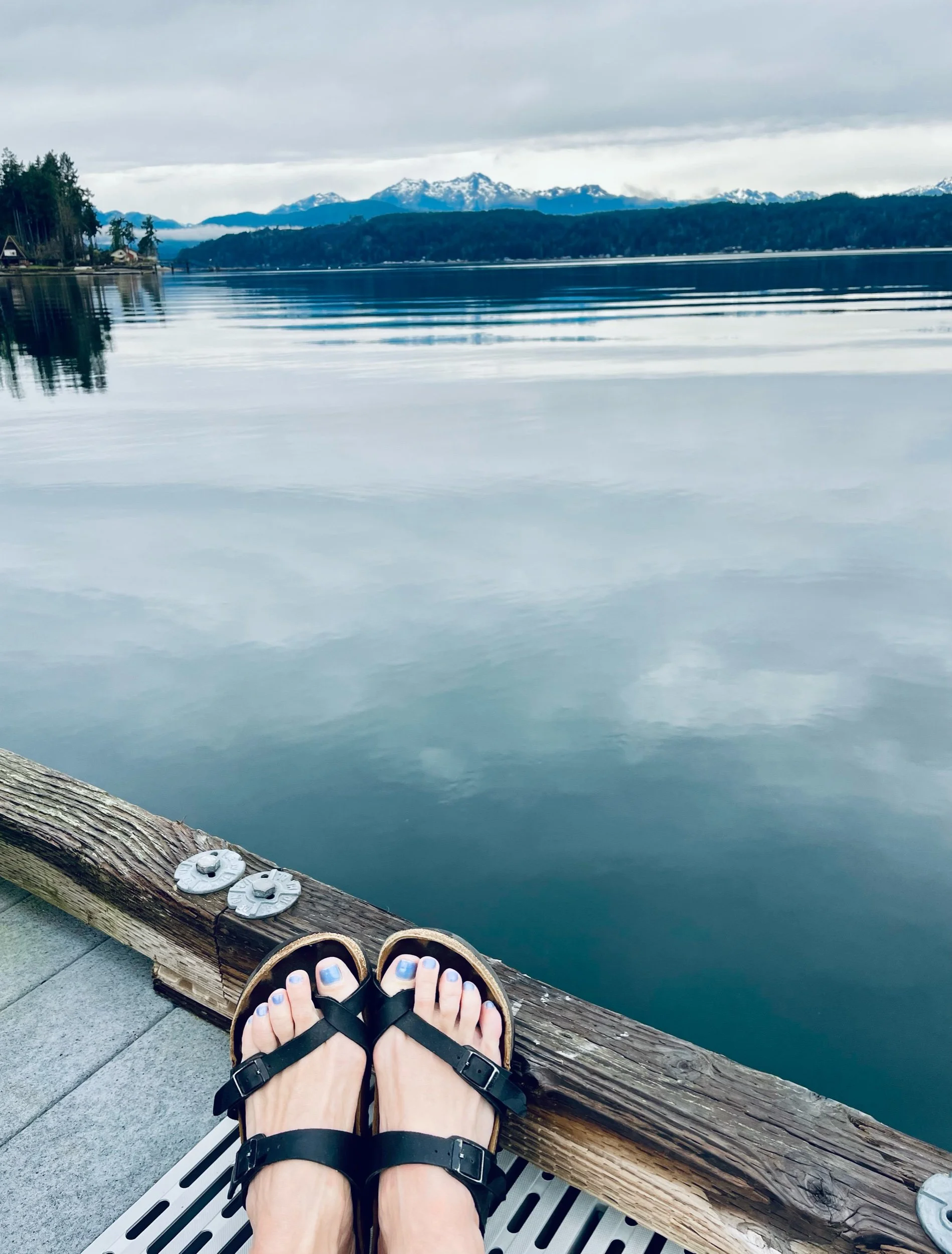 View of a person sitting on a dock by a lake, with feet in black sandals, painted toenails, and a wooden railing in the foreground. The lake has calm water, reflecting the cloudy sky, with mountains and trees in the background.