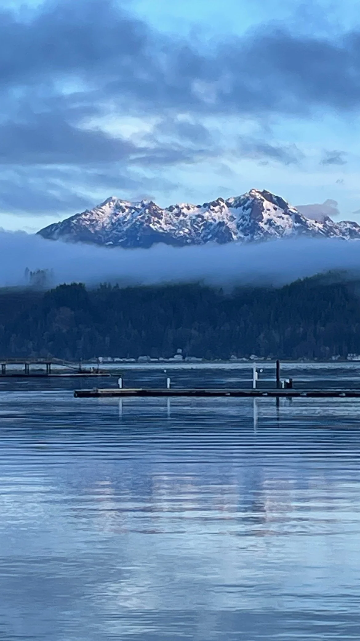 Snow-capped mountains under a cloudy sky across a body of water with docks or piers in the foreground.