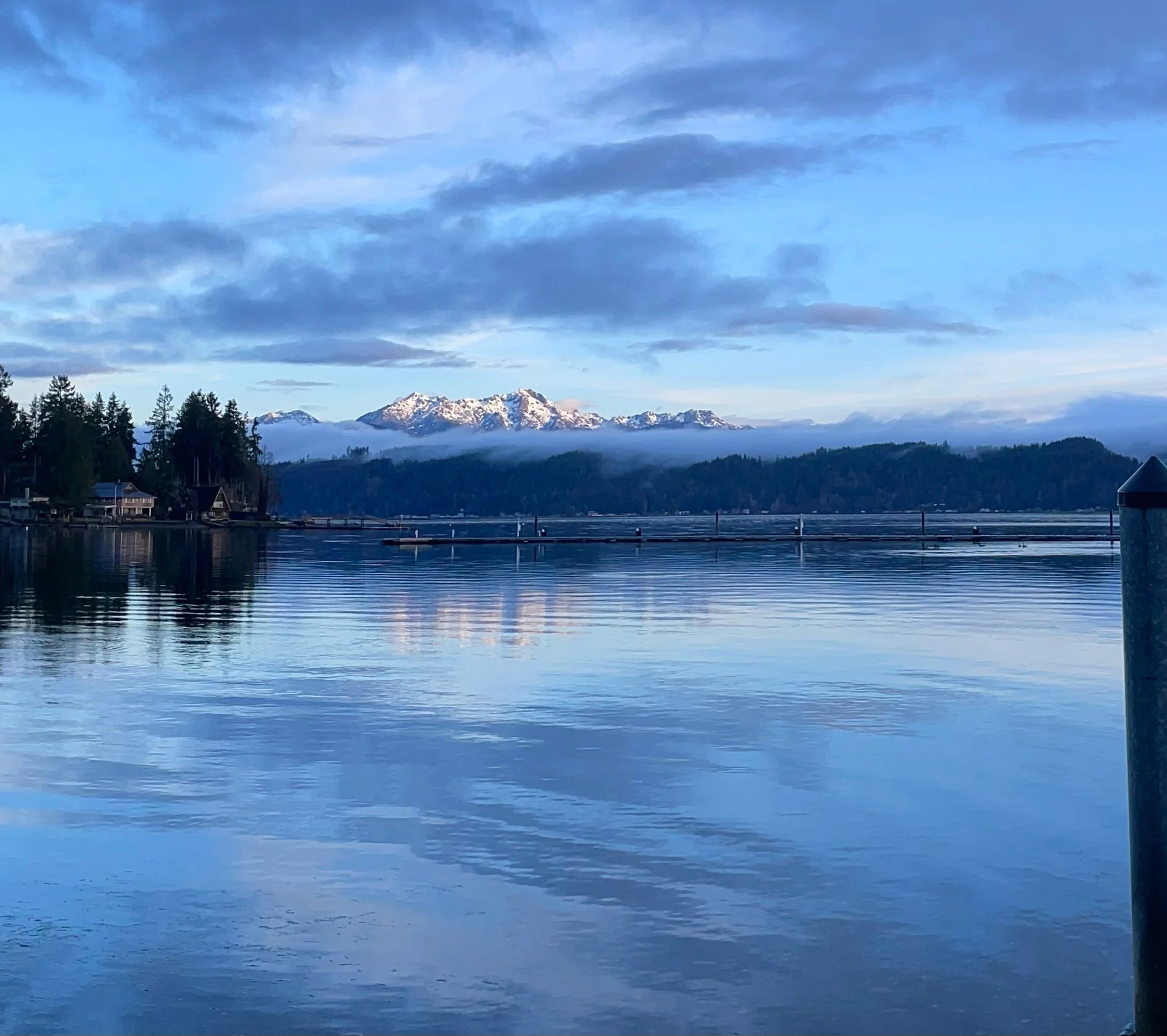 A large body of water reflecting the cloudy sky above, with snow-capped mountains in the distance and a small building or boathouse near the water's edge. Tall trees are visible along the shoreline.