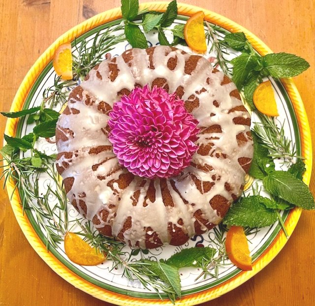 Ring-shaped cake topped with white icing and a pink flower in the center, garnished with mint leaves, orange wedges, and sprigs of herbs on a decorative plate.