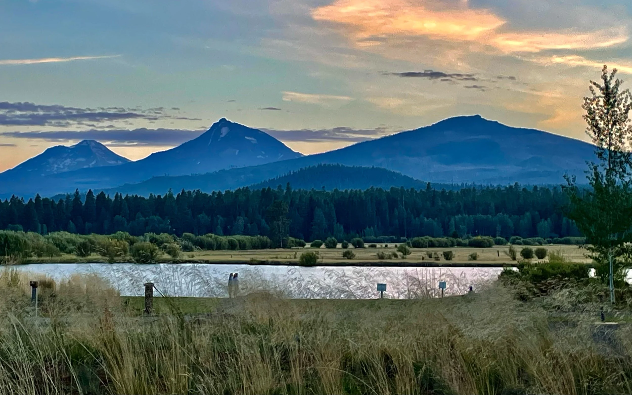 Scenic view of mountains with two prominent peaks, forested slopes, a body of water, and a sky with clouds during sunset.
