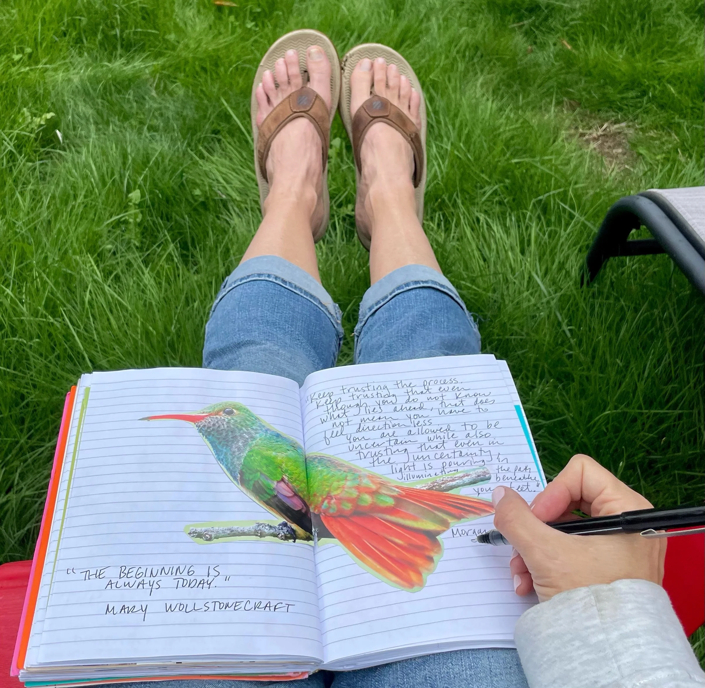 A person's legs wearing sandals with a notepad on their lap, outdoors on green grass, with a handwritten note about trusting and believing in oneself.