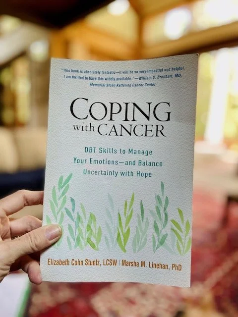 A person holding a book titled "Coping with Cancer: DBT Skills to Manage Your Emotions and Balance Uncertainty with Hope". The cover features green leafy illustrations and quotes from a medical professional.