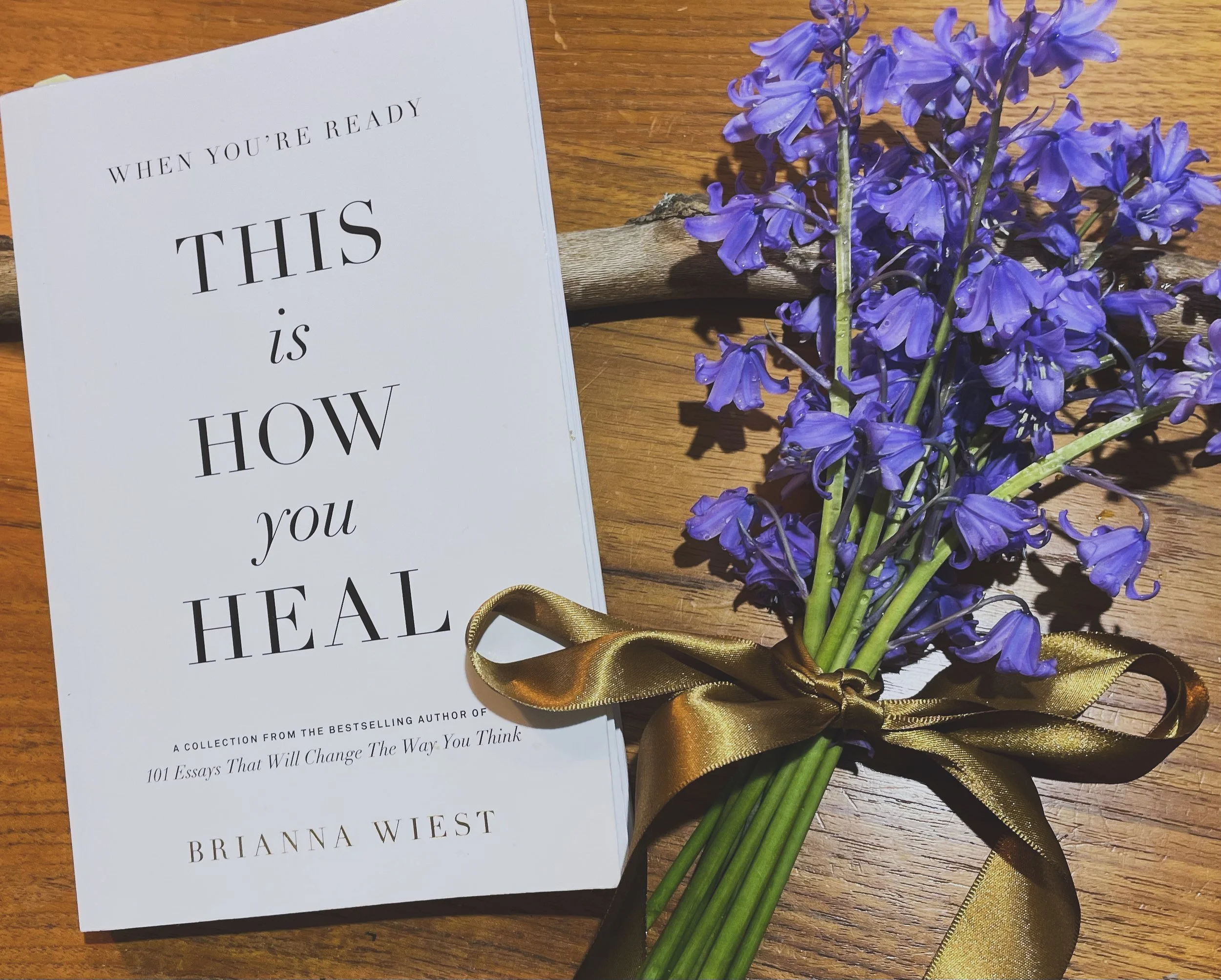 A close-up view of a white book titled 'When You're Ready This Is How You Heal' by Rianna West, placed on a wooden surface, next to a bouquet of purple flowers tied with a gold ribbon.