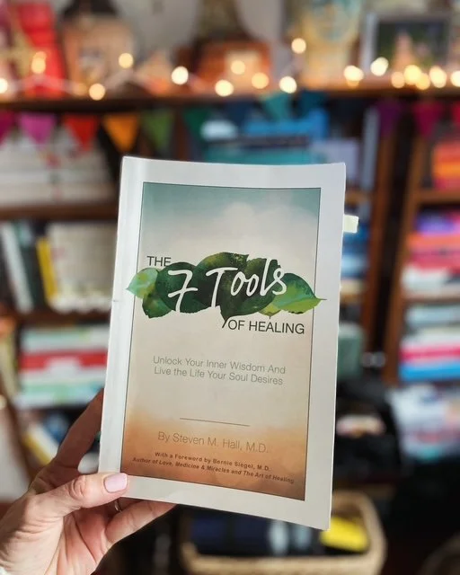 Person holding a book titled '7 Tools of Healing' in front of bookshelves decorated with string lights.