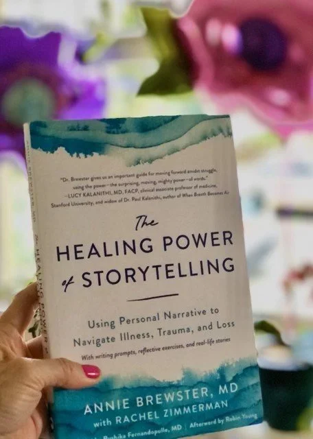 A person holding a book titled "The Healing Power of Storytelling" by Annie Brewster, MD, with purple flowers in the background.