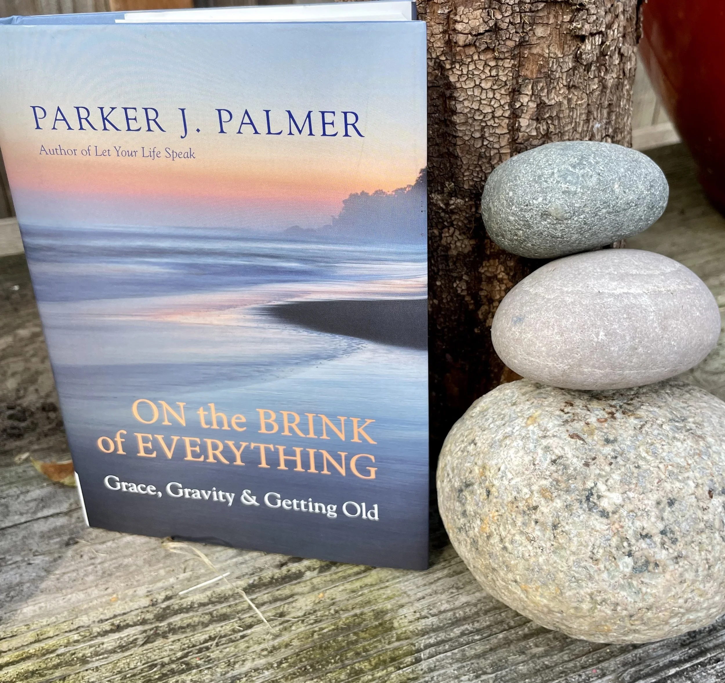 A book titled 'On the Brink of Everything' by Parker J. Palmer is placed on a weathered wooden surface next to some smooth gray stones and a tree trunk.