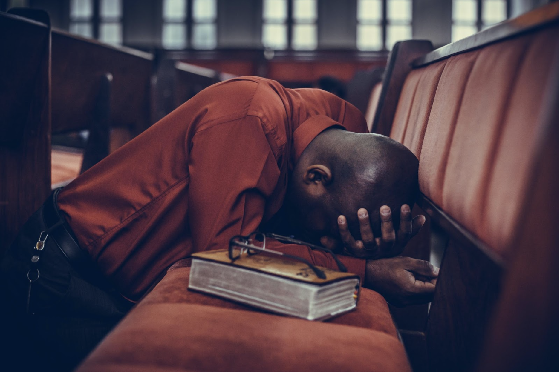 Black man praying in church pew appearing distraught with his hand on his head and experiencing some of the effects of religious trauma.