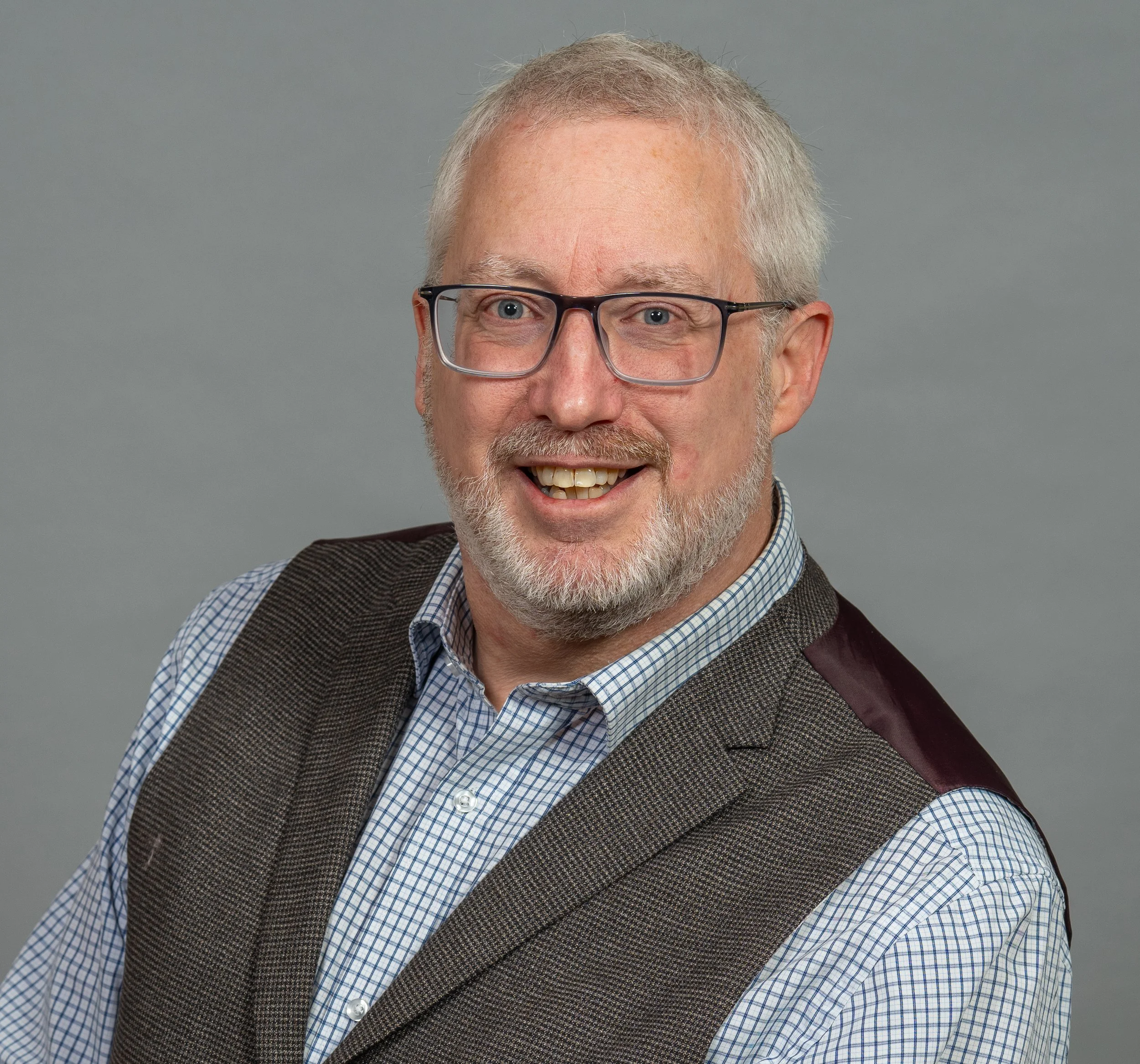 Portrait of a middle-aged man with a beard and glasses, wearing a checkered shirt and a brown vest, smiling against a plain gray background.