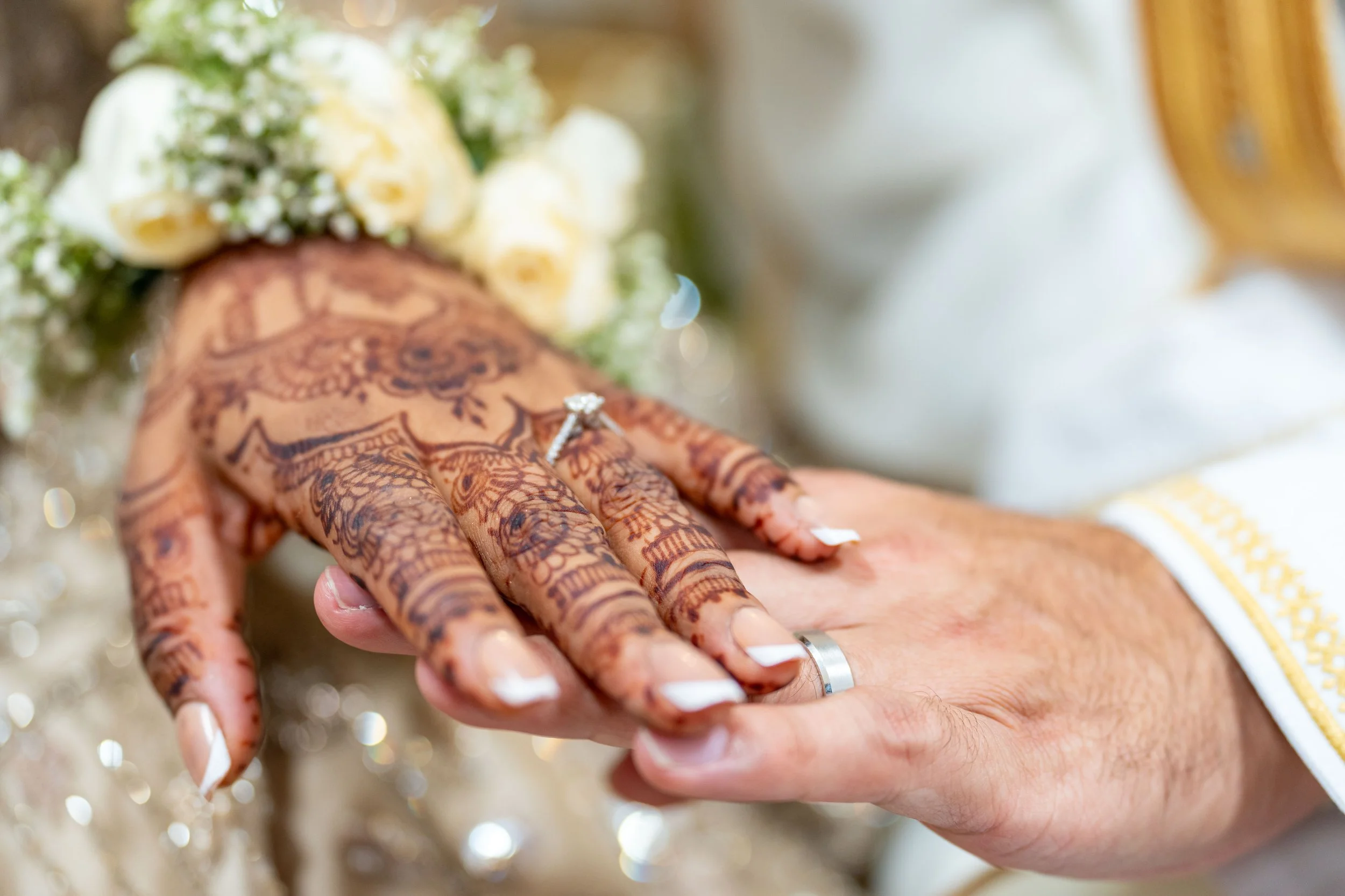 Close-up of a bride's hand with intricate henna designs, adorned with wedding rings, holding a bouquet of white roses with baby's breath, during a wedding ceremony.