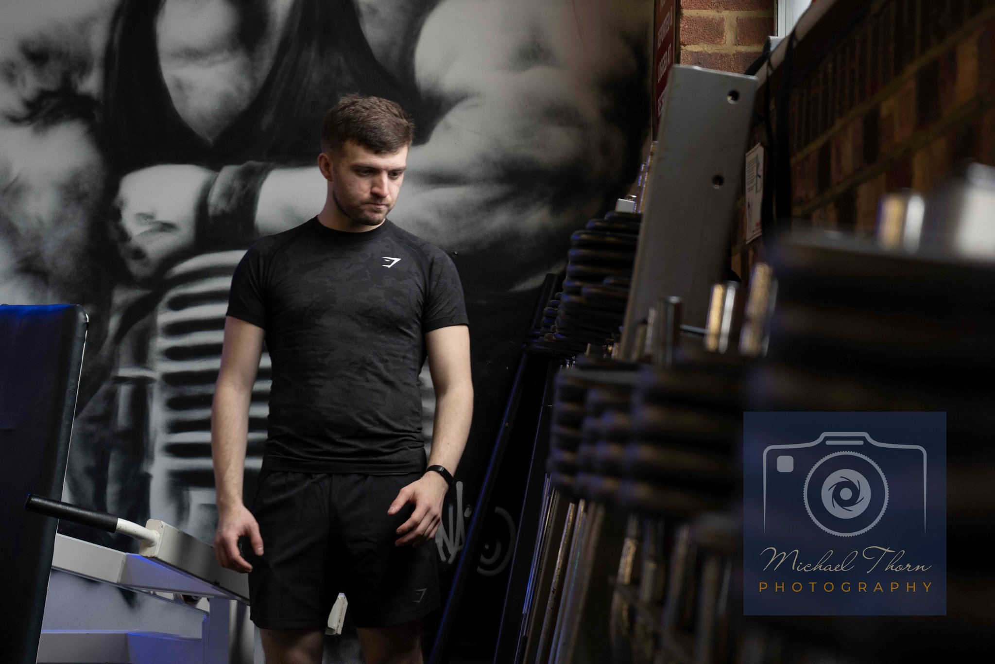 A young man in black athletic clothing looking down in a gym. In the background, there is a black and white mural of a person wearing goggles and a mask. To his right, there are shelves with stacked items and a coaching or workout station.
