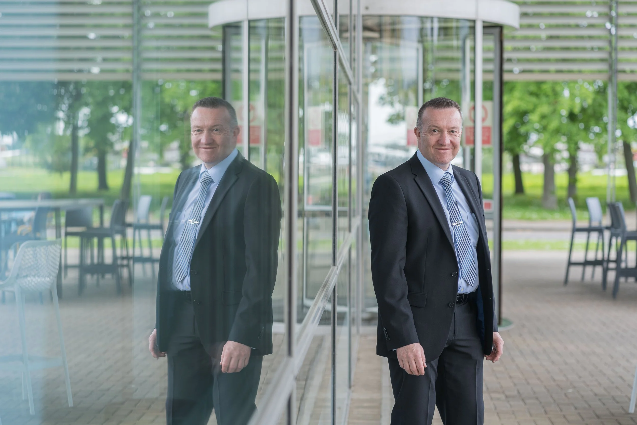 A middle-aged man in a suit stands outside, leaning against a glass wall with his reflection visible. He is dressed in a dark suit with a light blue shirt and striped tie, smiling at the camera with a park and trees in the background.