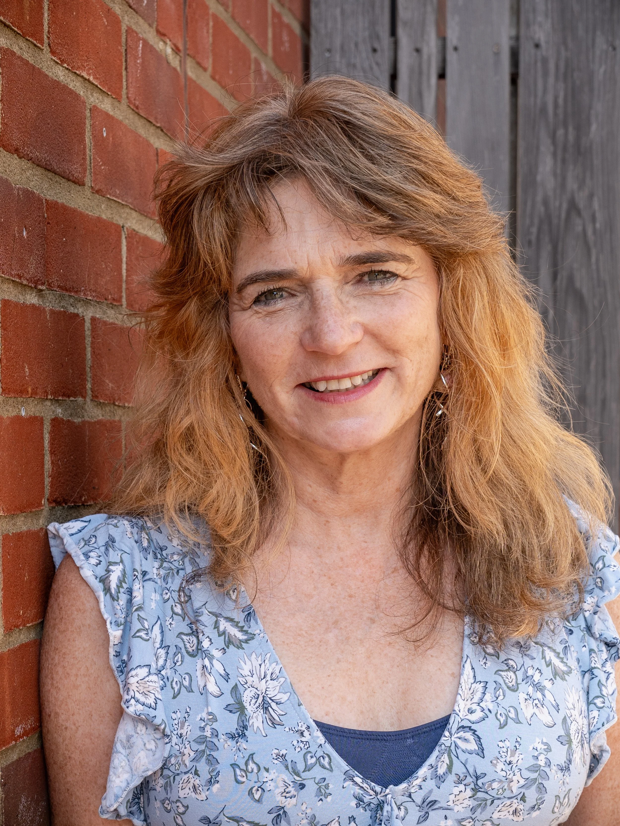 A smiling woman with reddish-brown hair leaning against a brick wall.