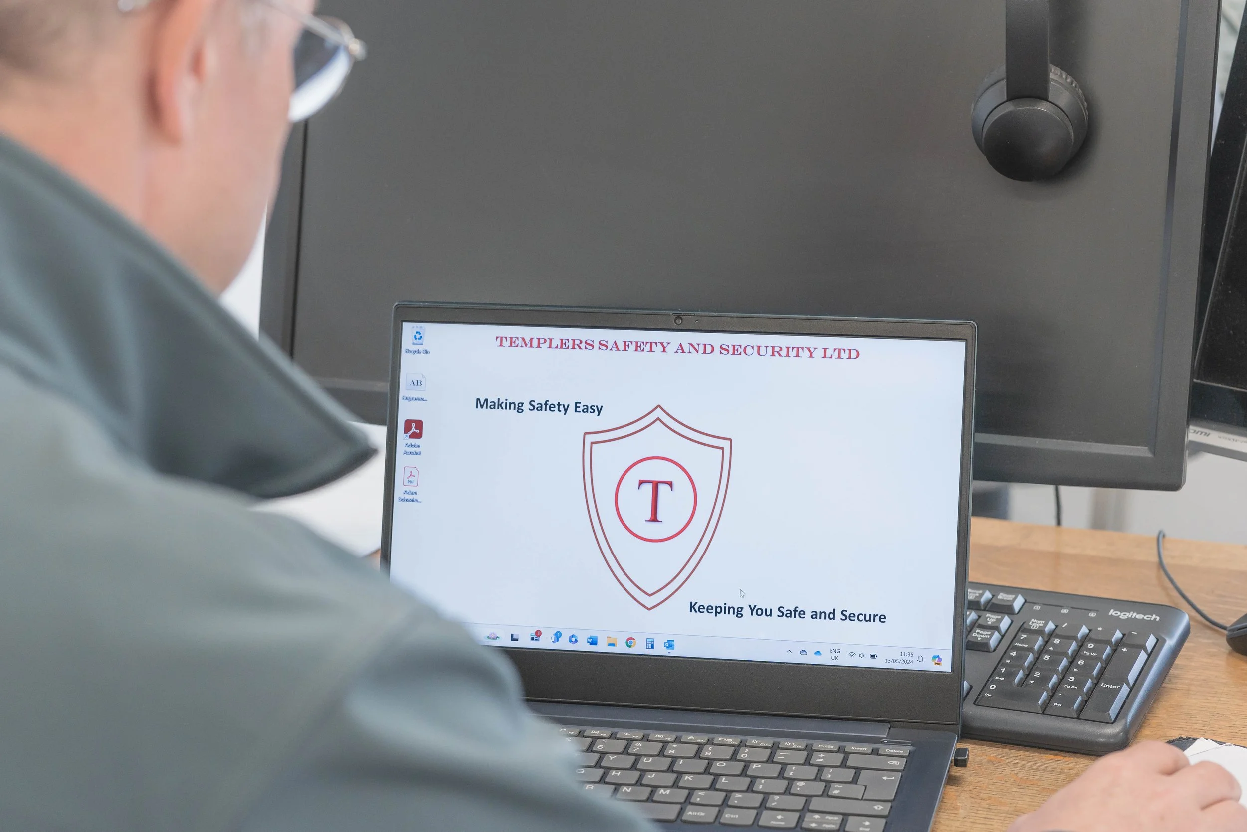 A person working at a computer with a monitor displaying a safety and security company's website, showing a shield with a T inside and the text 'Making Safety Easy' and 'Keeping You Safe and Secure.'