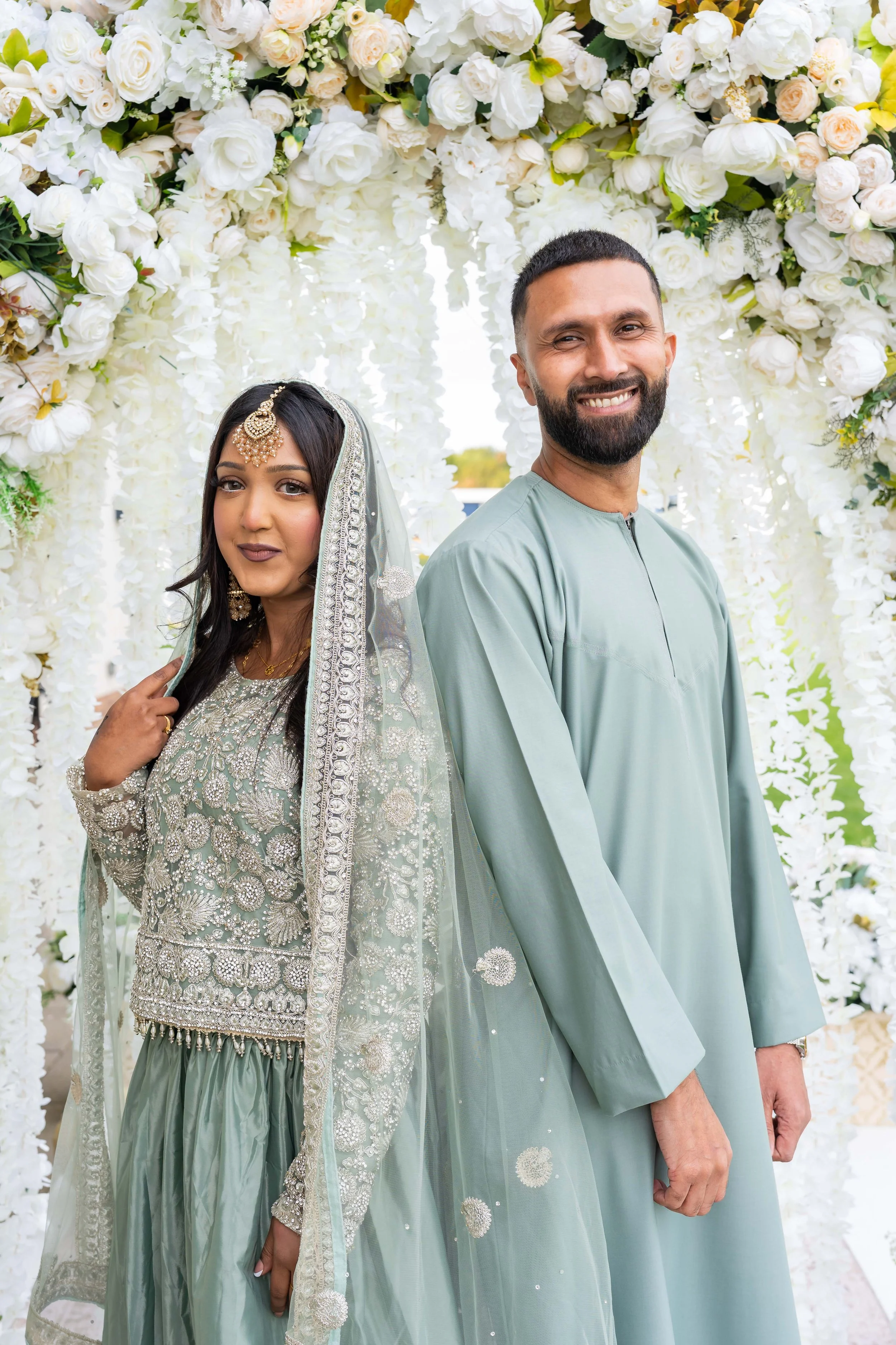 A bride and groom at their wedding, standing under a floral arch made of white and cream flowers.