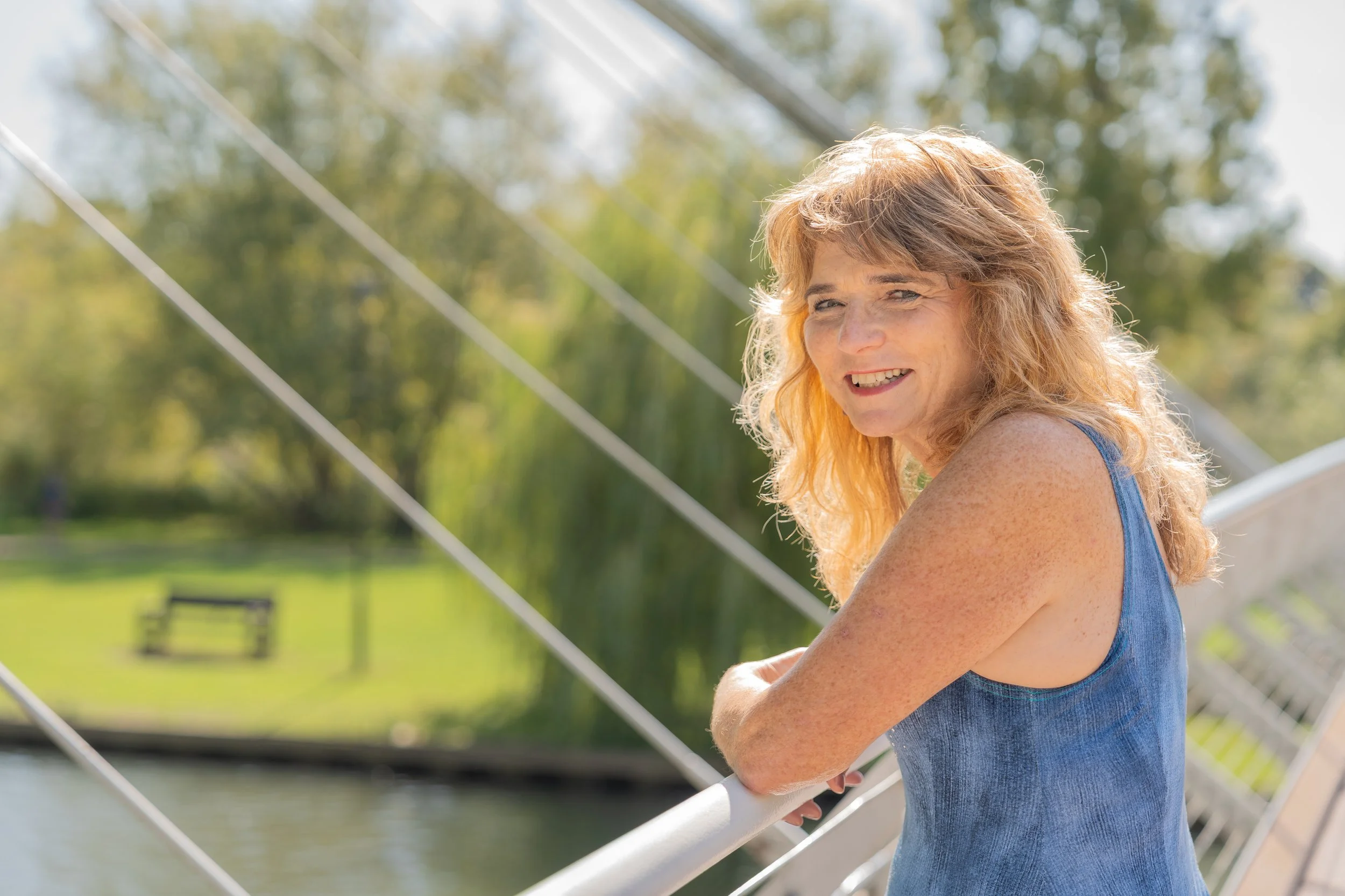 A woman with curly red hair wearing a sleeveless blue denim top, standing outdoors on a bridge, smiling and looking towards the camera, with a park featuring green grass, trees, and a bench in the background.