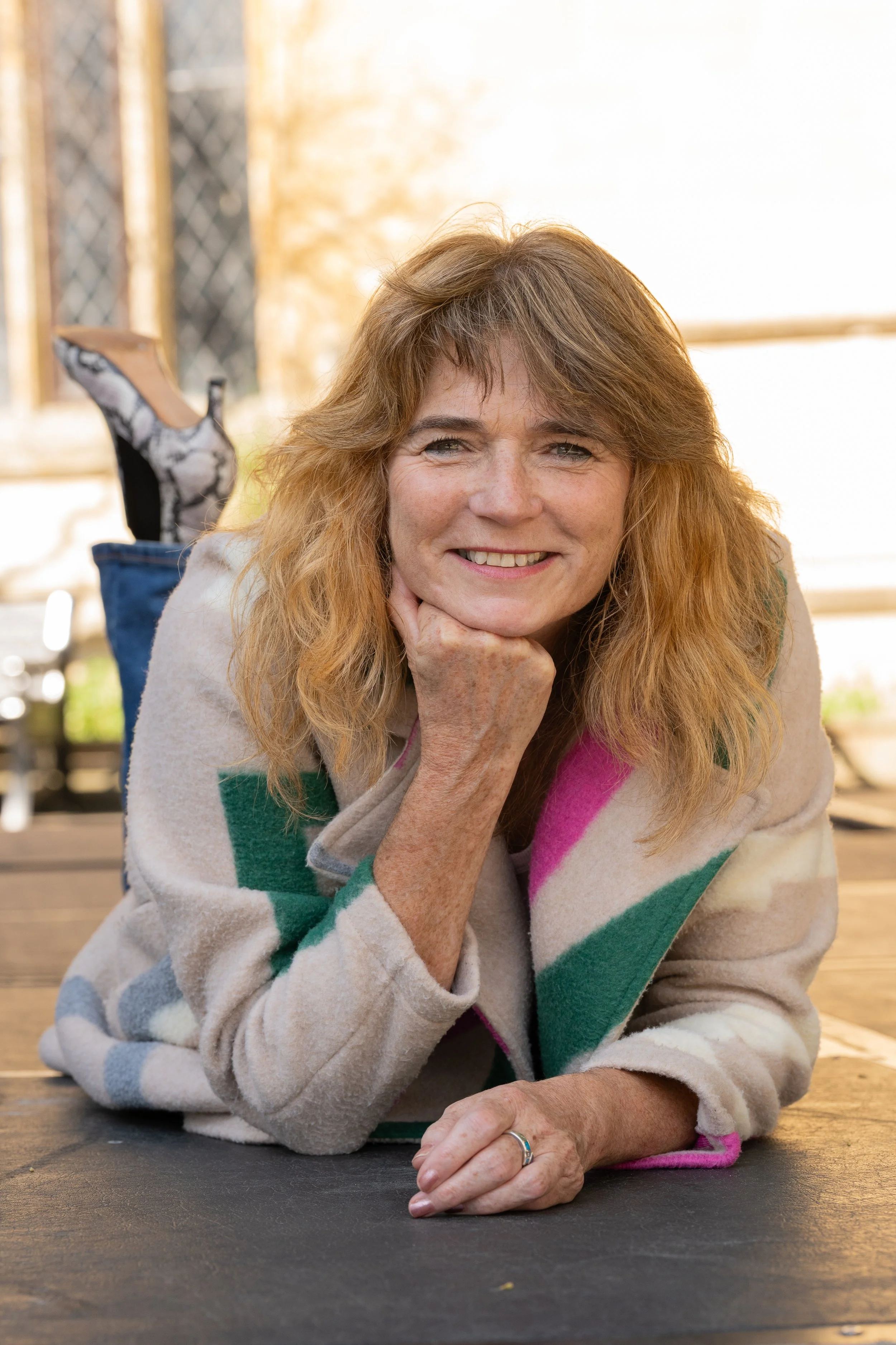 A woman with curly red hair laying on her stomach, resting her chin on her hand, smiling at the camera, wearing a colorful patterned coat, in an outdoor setting with a wooden surface and chains-link fence in the background.
