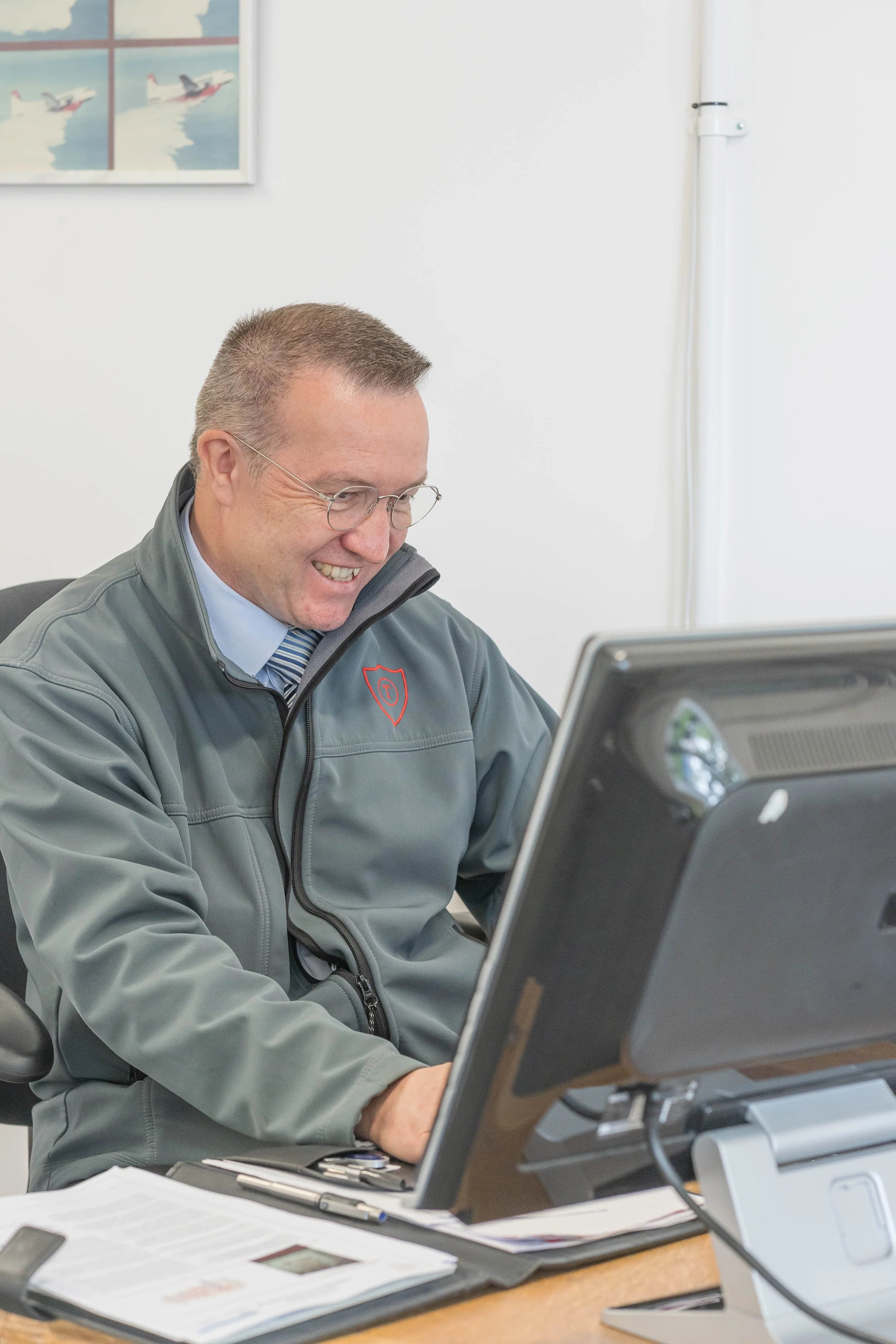 A man in a gray jacket and glasses smiling while working at a desk with a computer in an office.