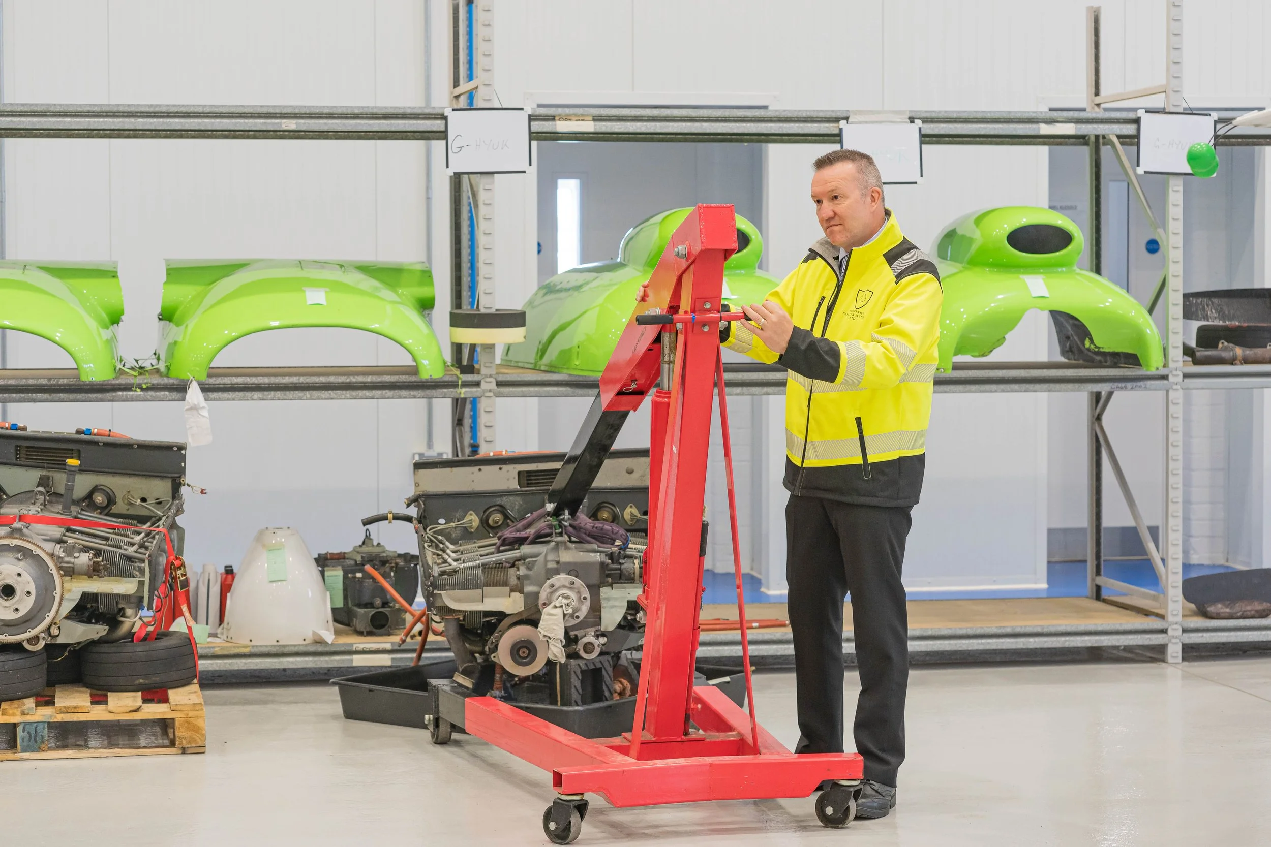 A man in a yellow and black jacket operating a red machine in a workshop. Behind him, green automotive body parts are stored on metal shelves, and various engine components are on the floor.