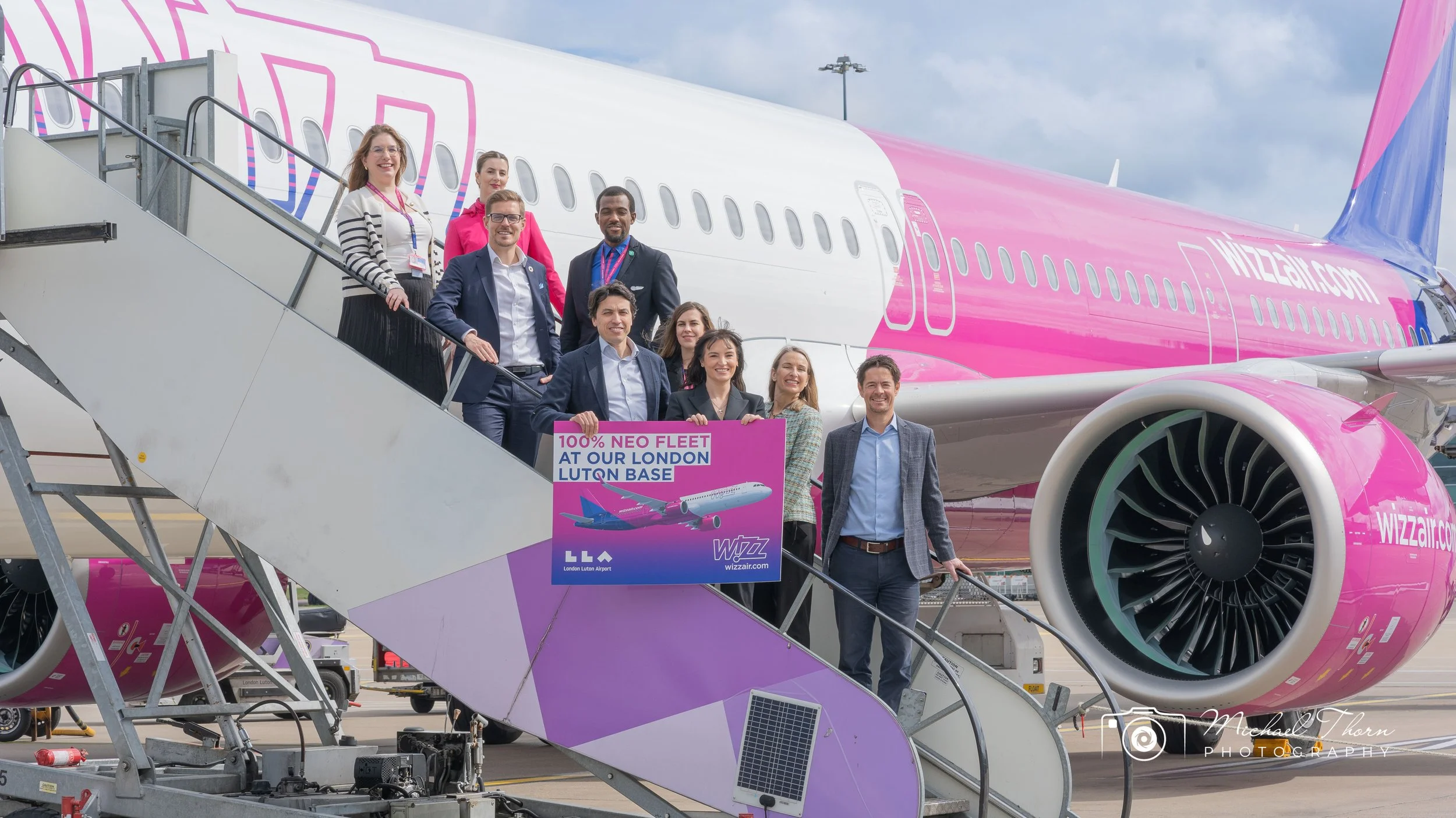 Group of people posing on the stairs of an airplane with pink and white branding at an airport, holding a sign that reads '100% Neo Fleet at Our London LUTQN Base' and a graphic of an airplane.