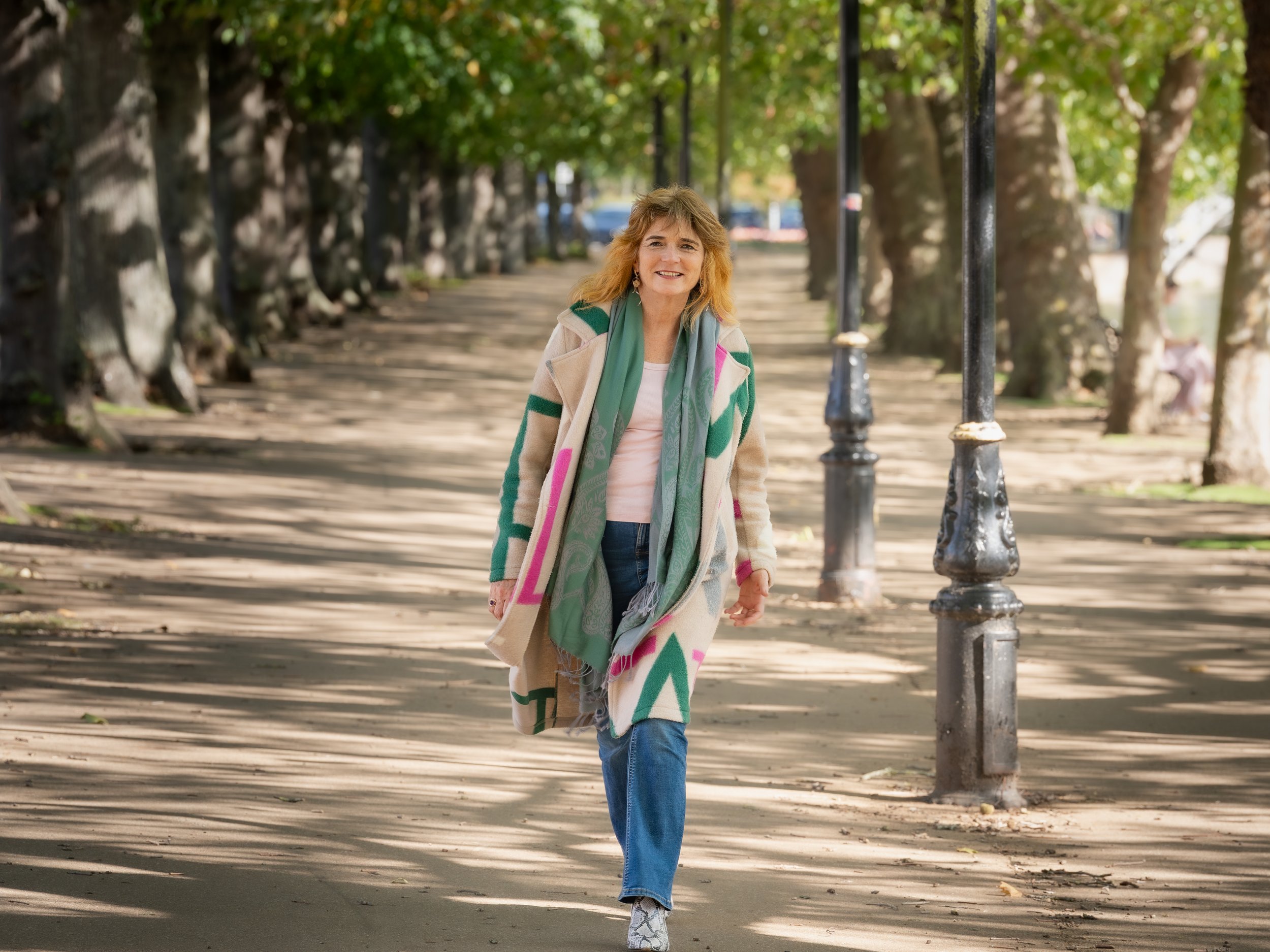 A woman with blonde hair walking along a tree-lined sidewalk, smiling, wearing a colorful coat and scarf.