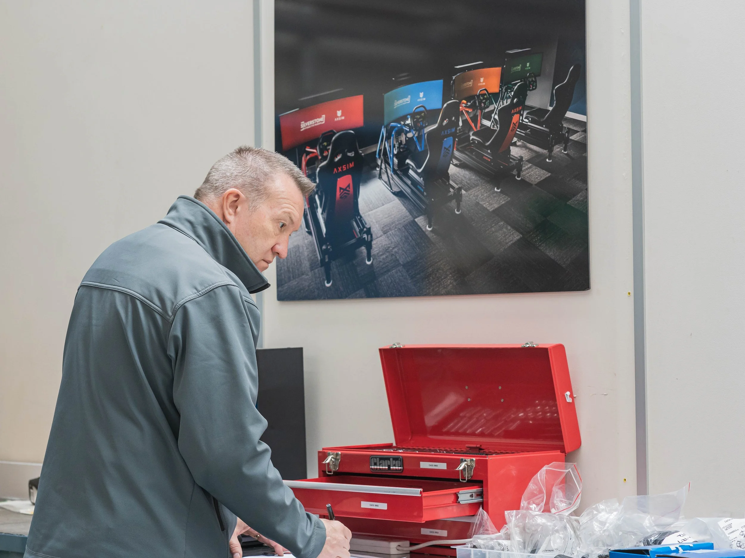 A man in a gray jacket working at a table with a red toolkit, in front of a wall with a large poster of racing simulator chairs and screens.