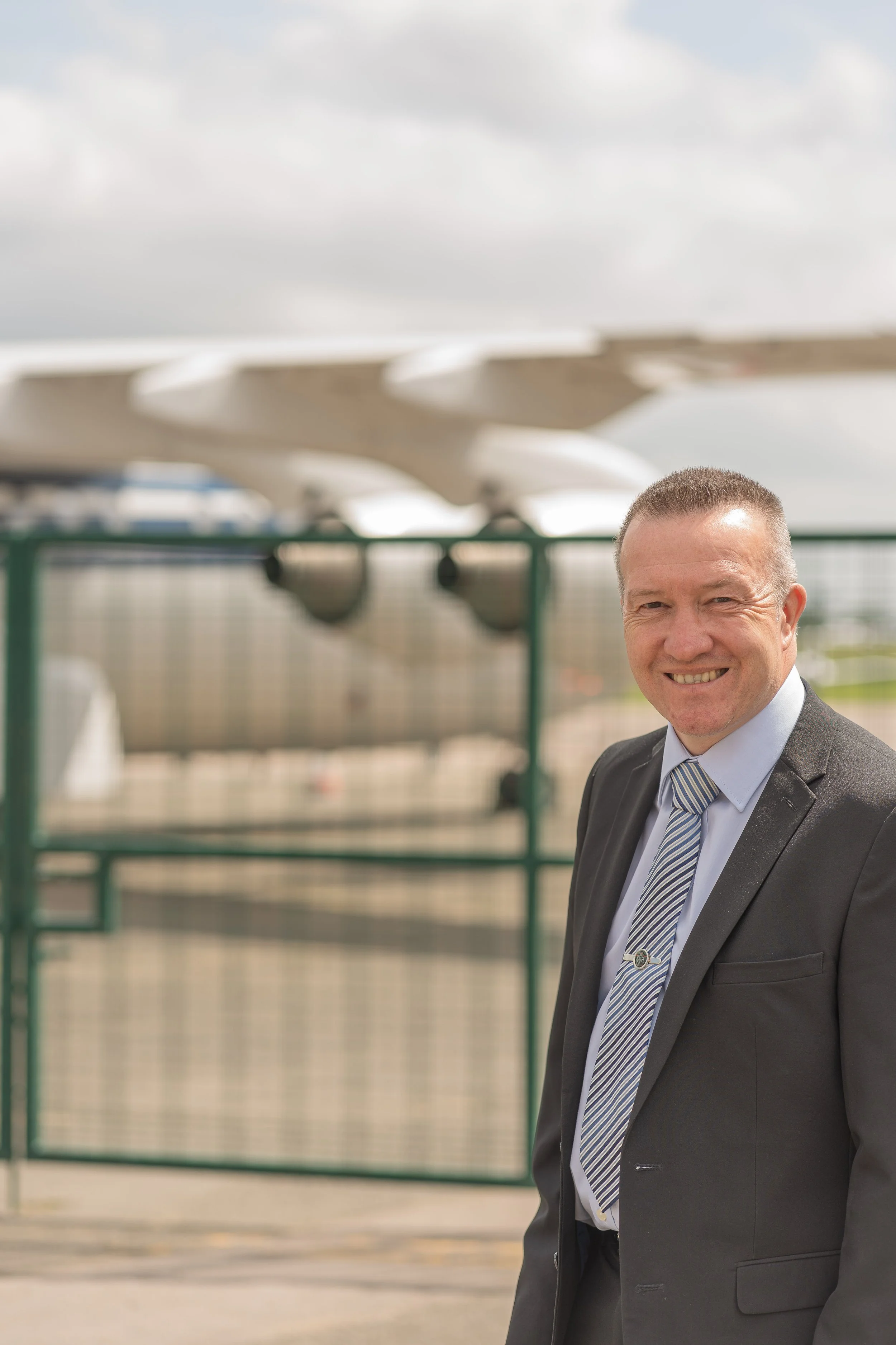 A man in a business suit standing outdoors in front of a fence, with an airplane tail in the background.
