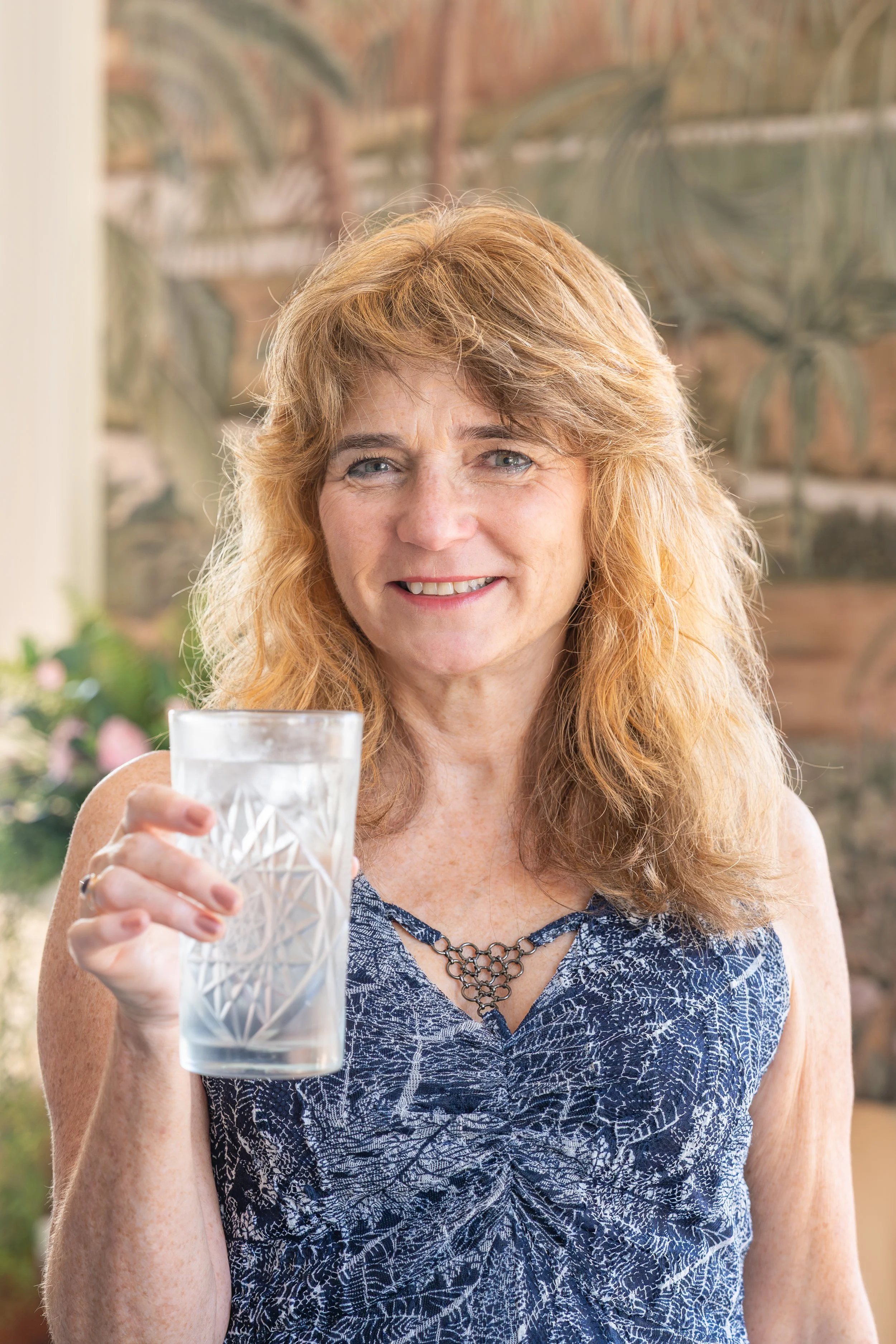A smiling woman with freckles and curly red hair holding a glass of water indoors, with a blurred background of plants and a brick wall.