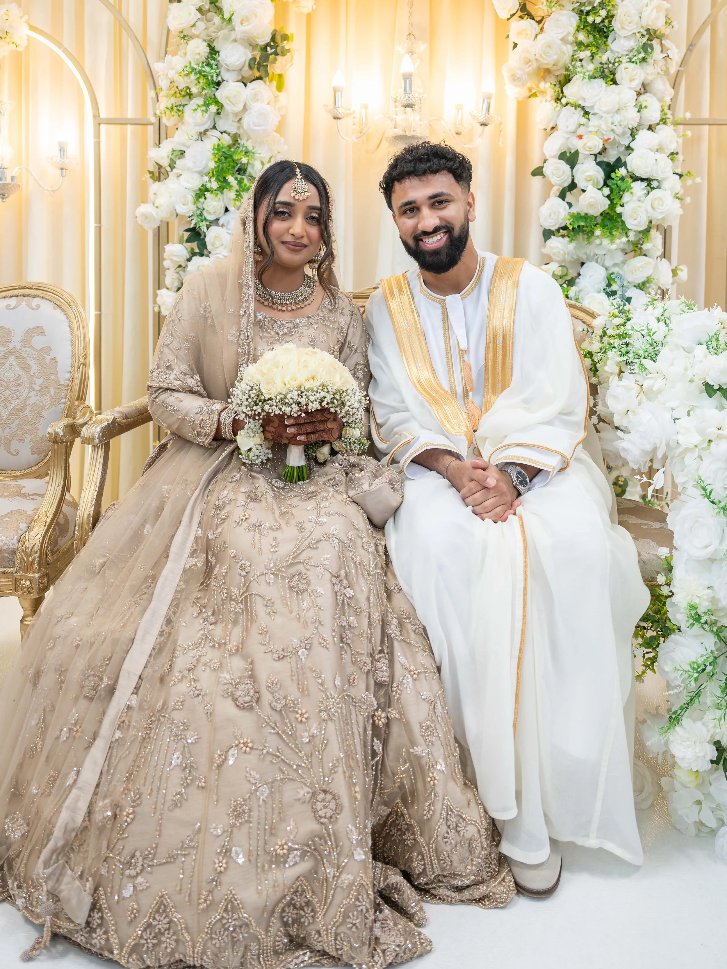 A newlywed couple seated on a decorated stage with a floral backdrop. The bride wears an ornate beige bridal gown and jewelry, holding a bouquet, and the groom wears a white traditional outfit with gold accents, both smiling at the camera.