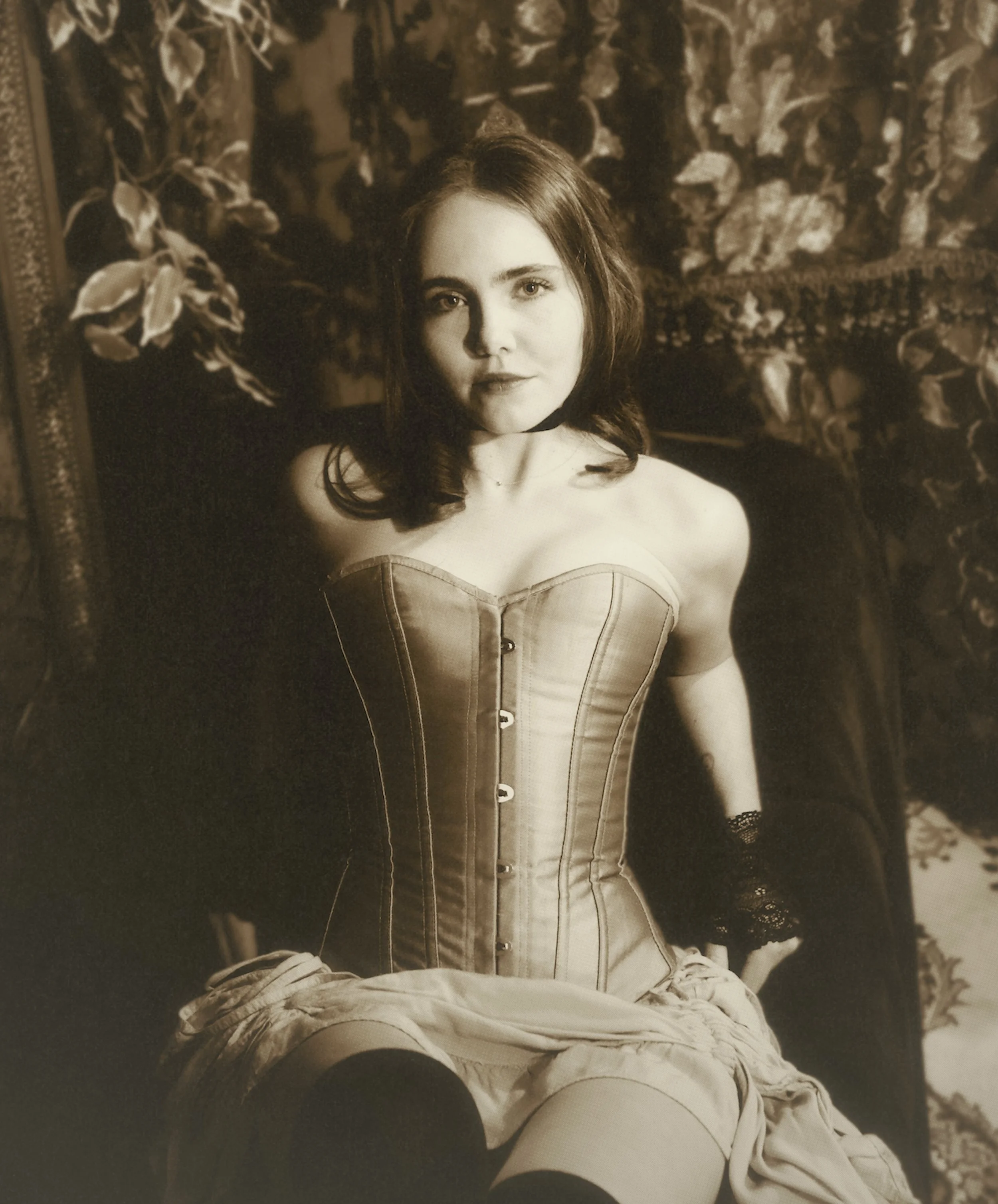 A young woman with dark hair, wearing a strapless corset and lace gloves, sitting in a vintage-inspired setting with floral wallpaper and ornate furniture.