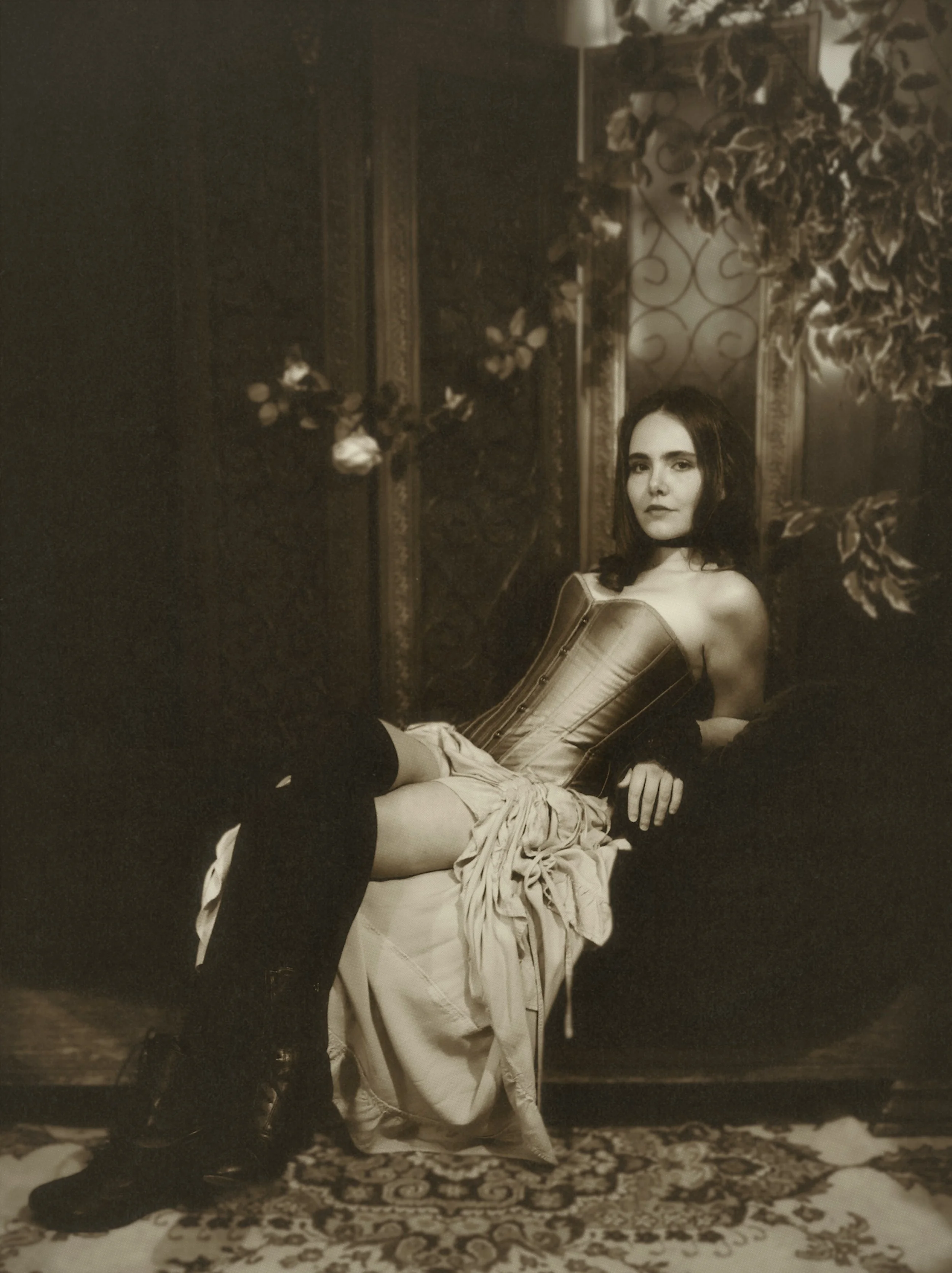 A woman with dark hair sitting on a sofa in a vintage-style room, wearing a corset and stockings, with a calm expression.