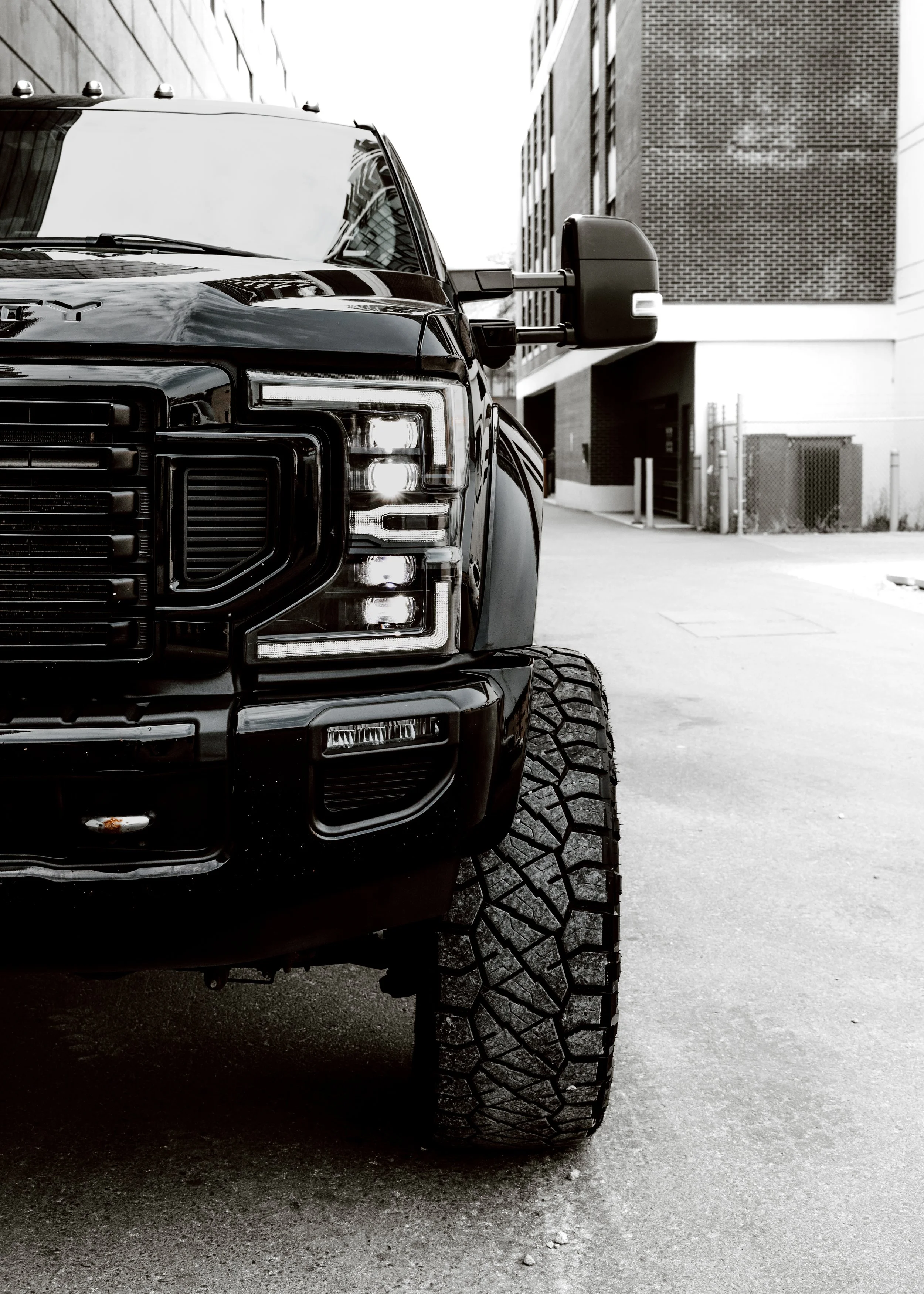 A black pickup truck with large tires parked in an urban alleyway, with modern buildings in the background.