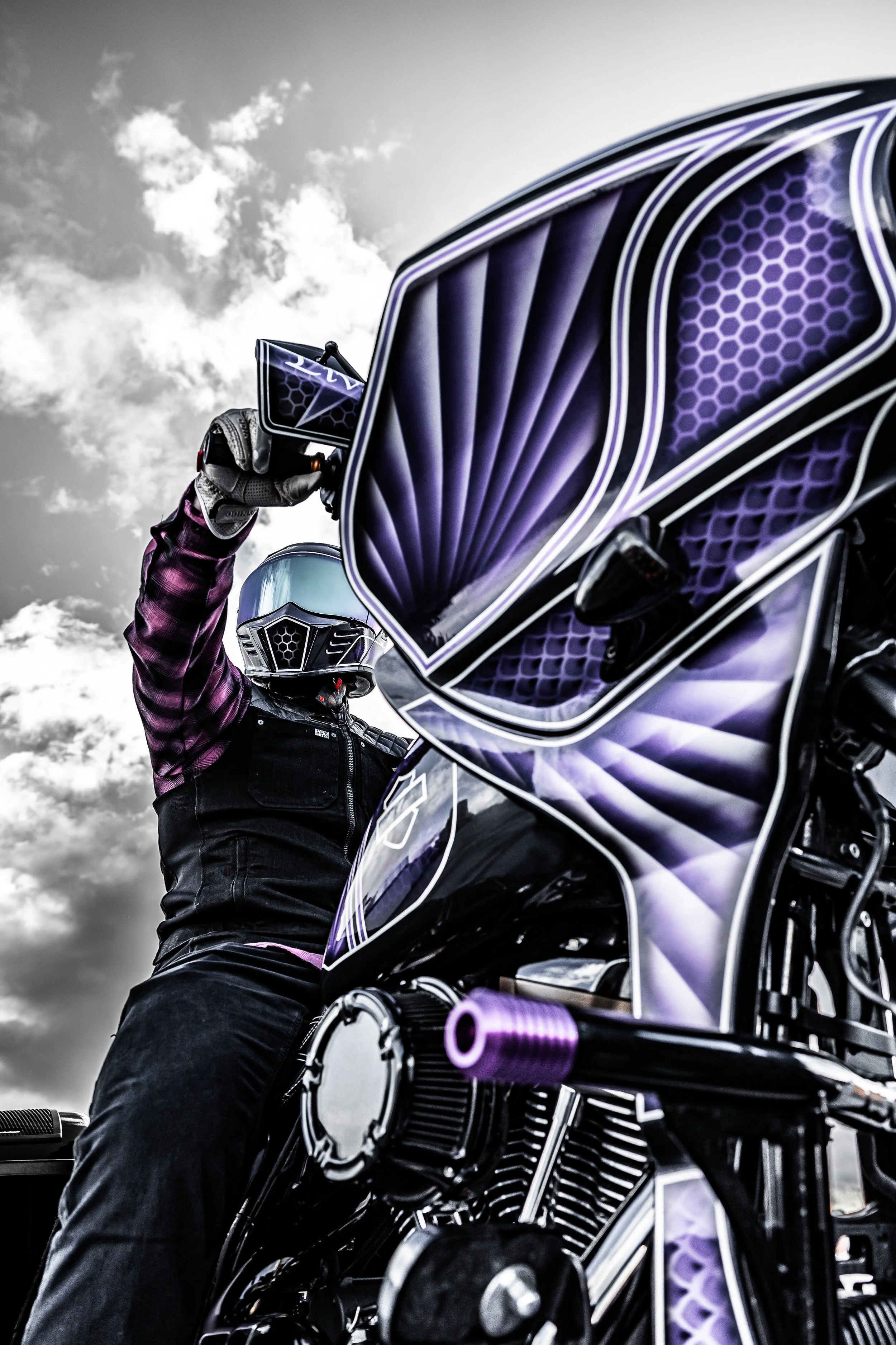 A person in a motorcycle helmet and black and purple riding gear adjusting a purple motorcycle helmet against a cloudy sky.