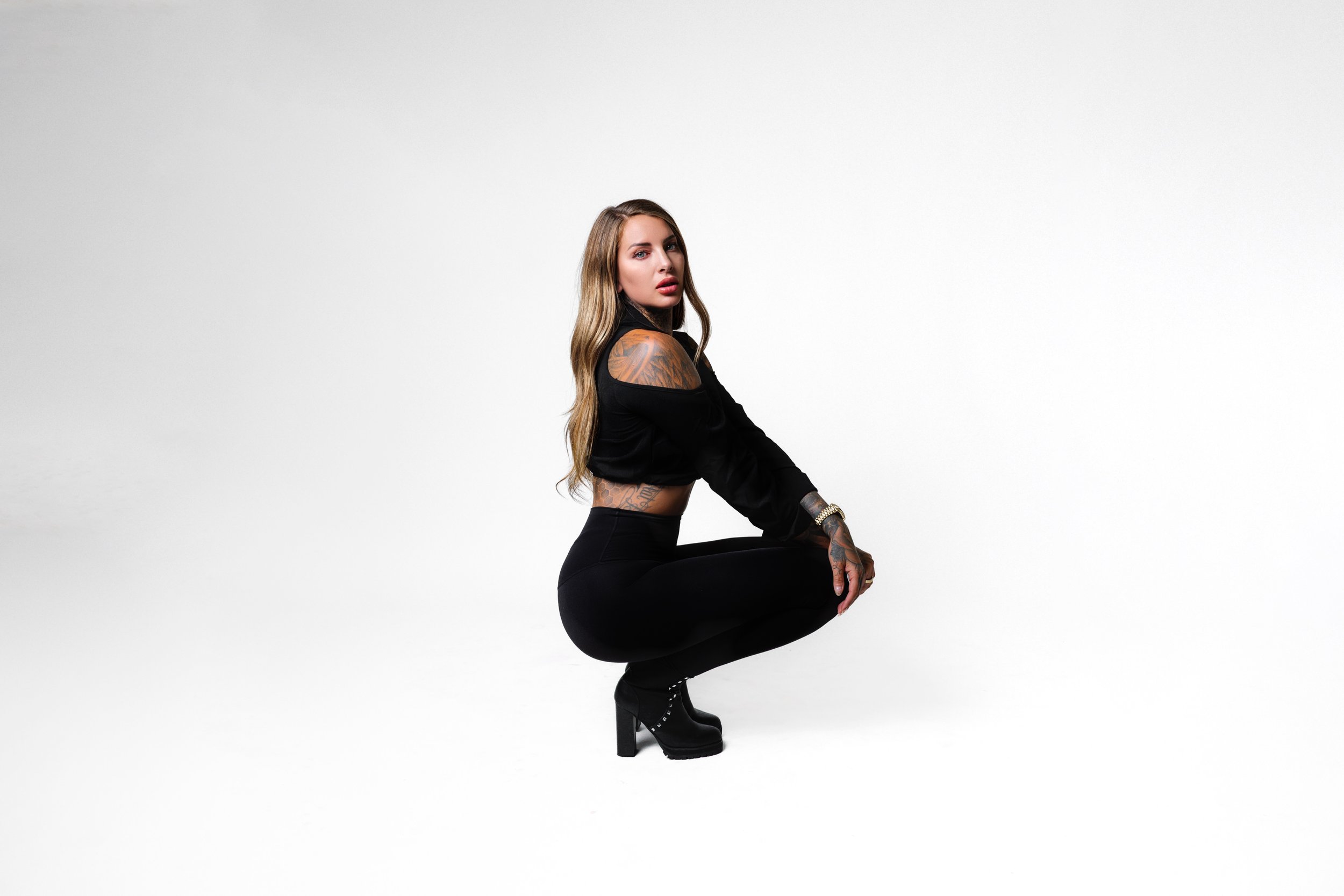 Woman with tattoos squatting in black outfit against white background.