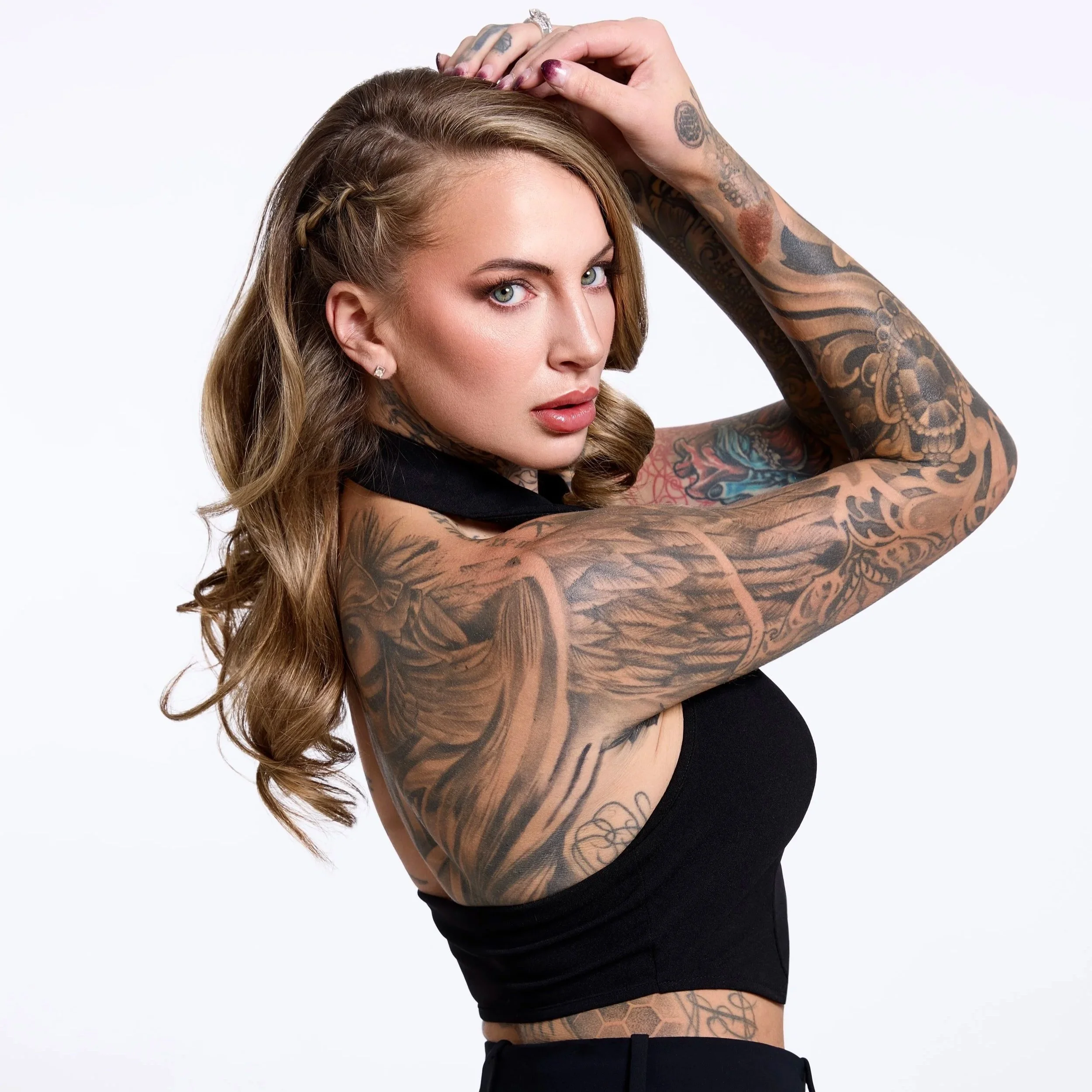 A woman with long, wavy hair and tattoos covering both arms, posed with her right arm raised and her hand resting on her head, wearing a black sleeveless top against a plain white background.