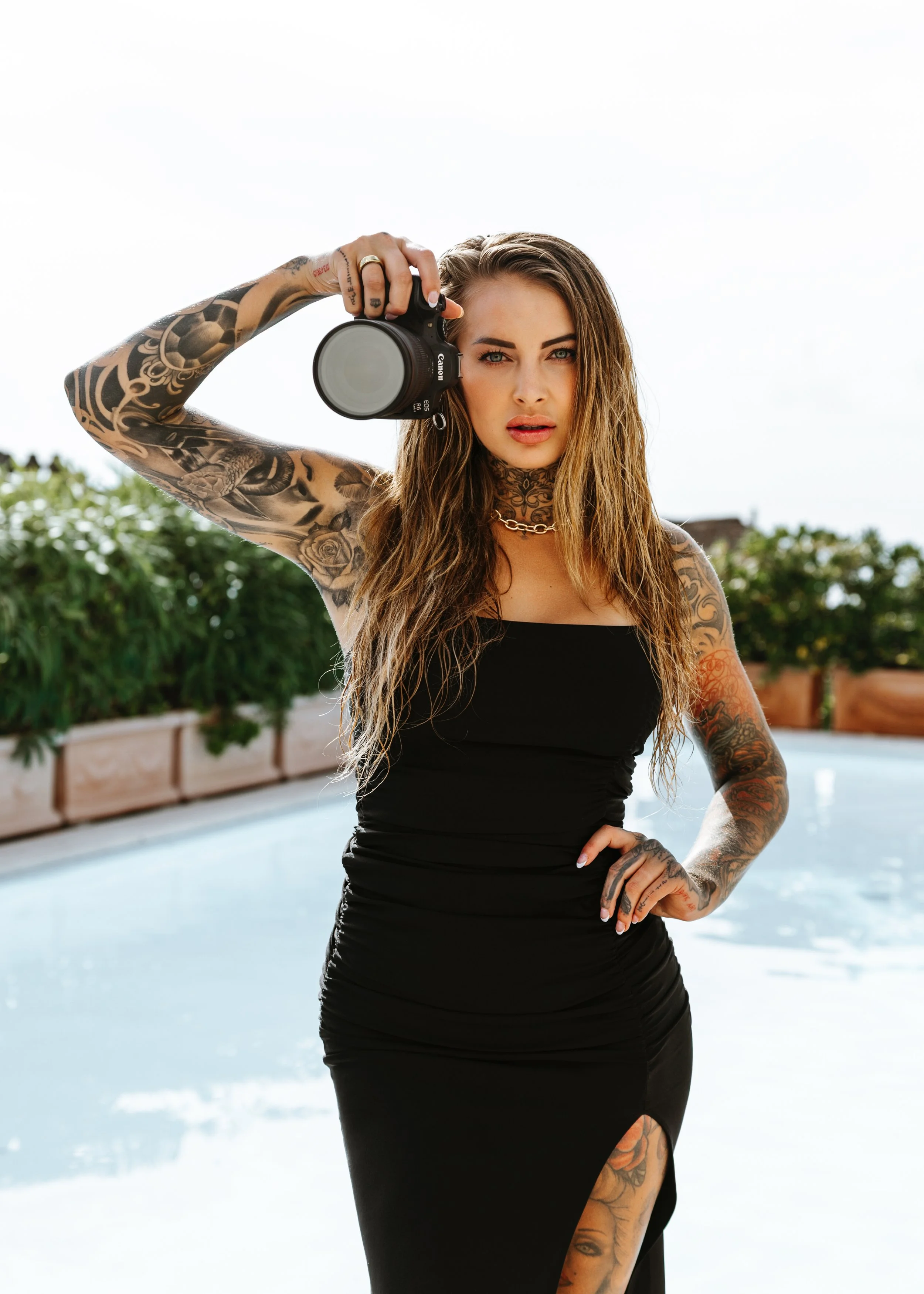 Woman with long wavy hair in a black dress, holding a camera to her eye near an outdoor pool, tattoos on her arms and neck, standing on a bright day.