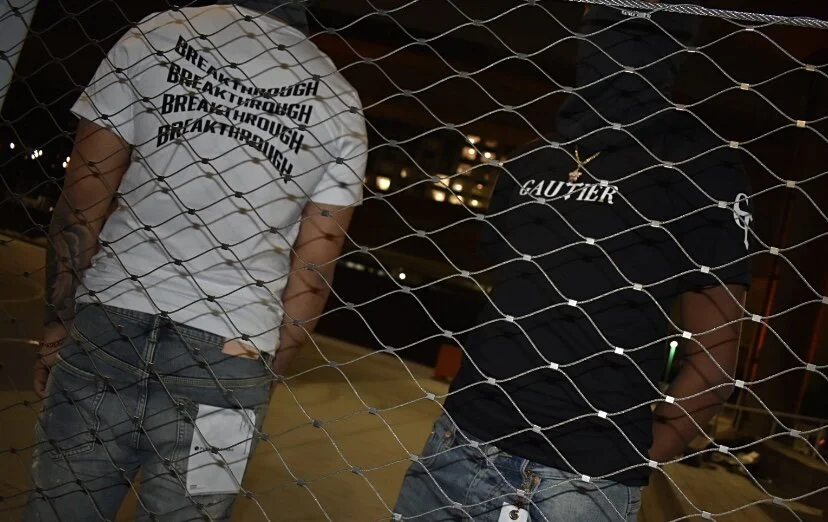 Two individuals standing behind a chain-link fence at night, with city lights in the background. One person wears a white shirt with 'BREAKTHROUGH' written multiple times, and the other wears a black shirt with 'GAUTIER' on it.