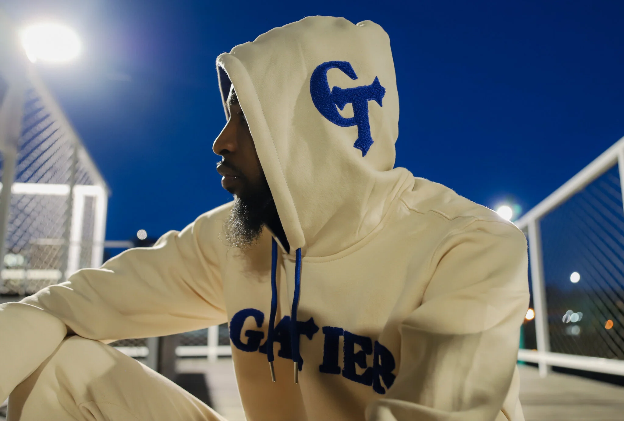 A person wearing a cream-colored hoodie with a blue design is sitting near a chain-link fence under a starry night sky, with bright stadium lights in the background.