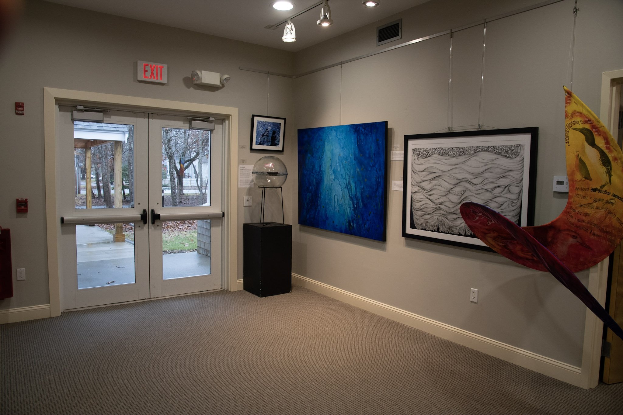  Several pieces of artwork hanging in the gallery, including "DCM: Party Zone" by Heather E. Stivison, "Turbulence" by Ellen Biegert, and "Marine Heatwaves" by Deb Ehrens 