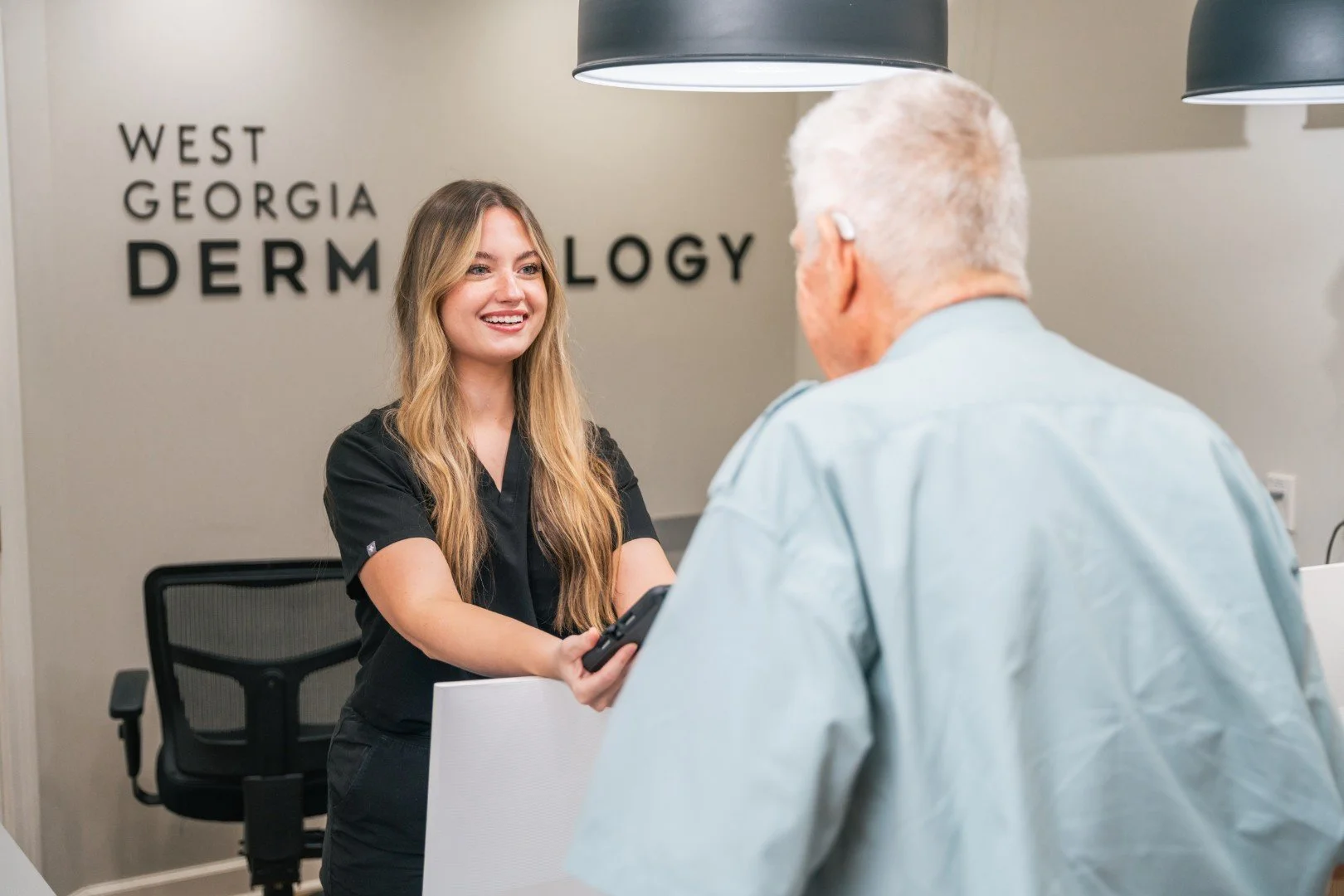 A young woman in medical scrubs handing a device to an older man in a medical office. The background has a sign that reads 'West Georgia Dermatology.'