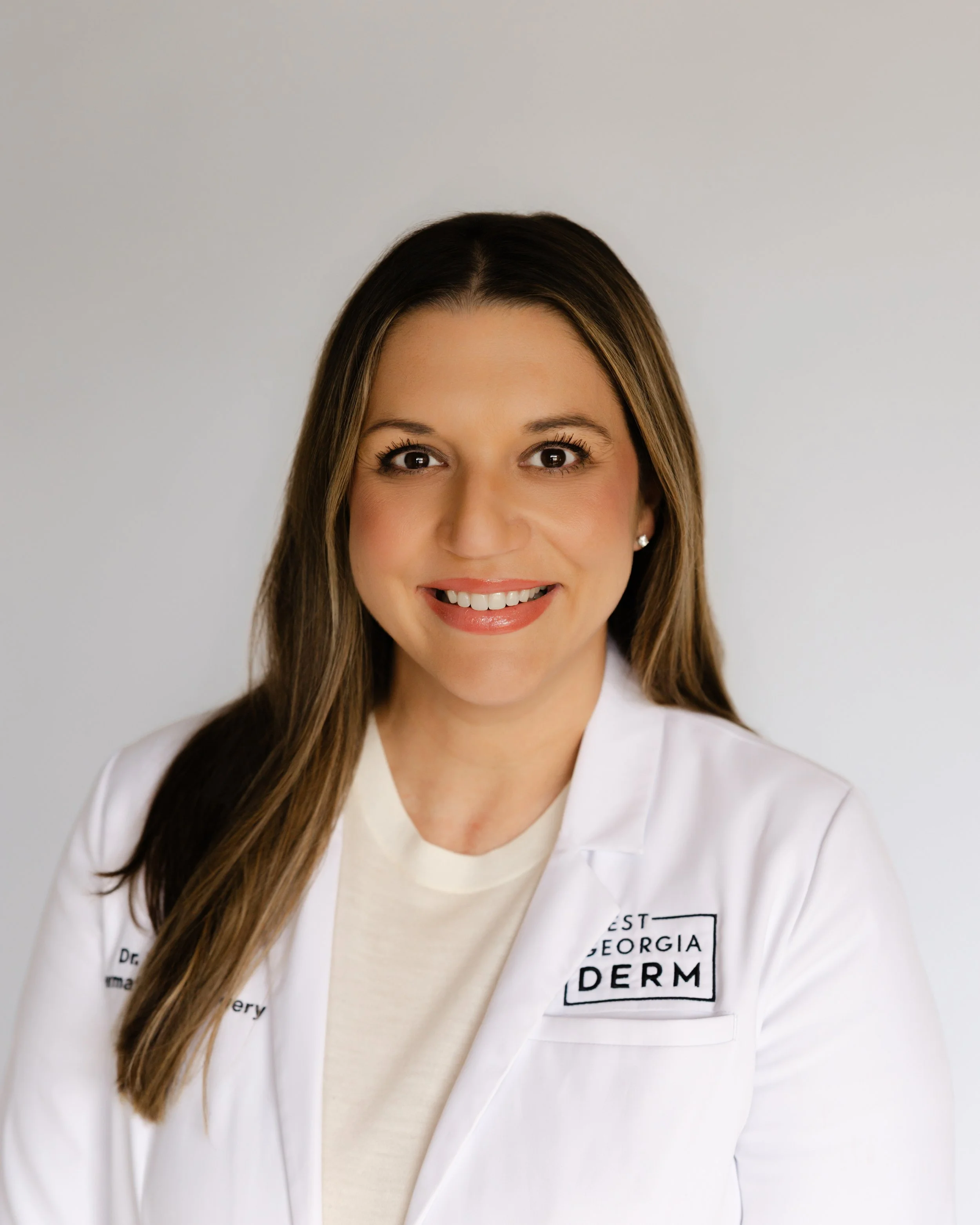Woman in a black shirt and white lab coat standing against a plain beige background.
