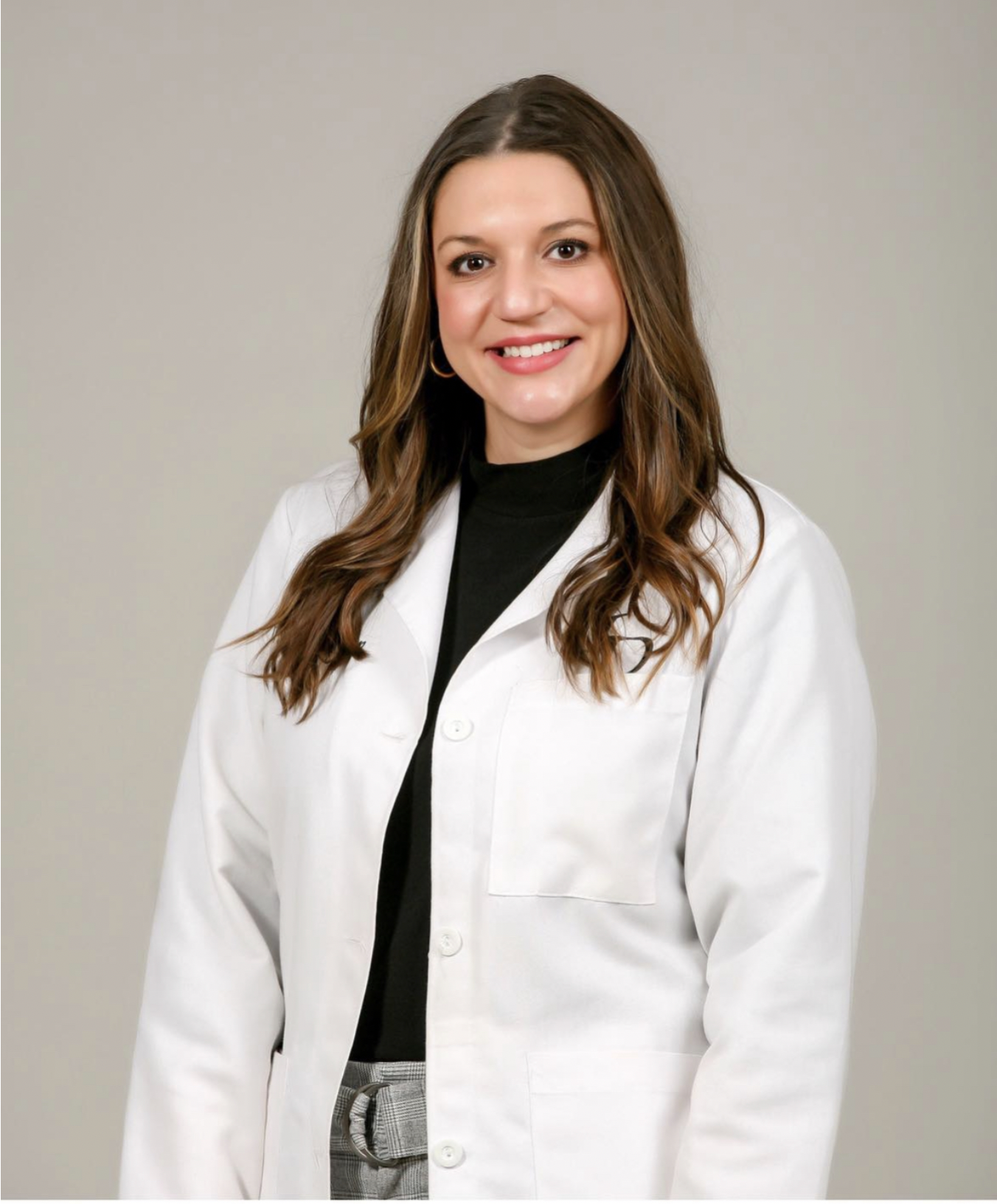 Woman in a black shirt and white lab coat standing against a plain beige background.